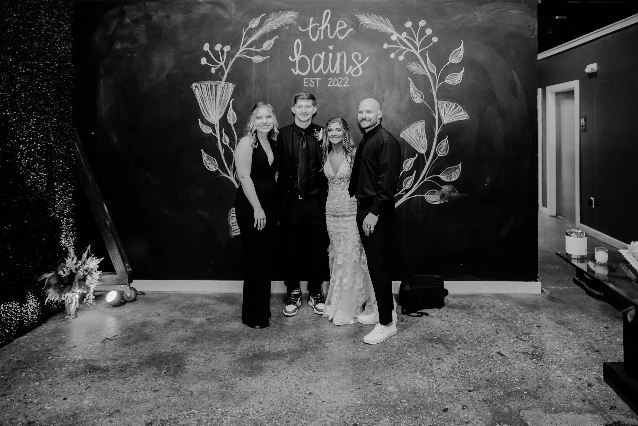 Four people standing in front of a decorative chalkboard with floral artwork and the words 'the bains, est 2022' written on it. All are dressed in formal attire, with the woman in a white lace wedding dress and the men in black suits, two with ties a