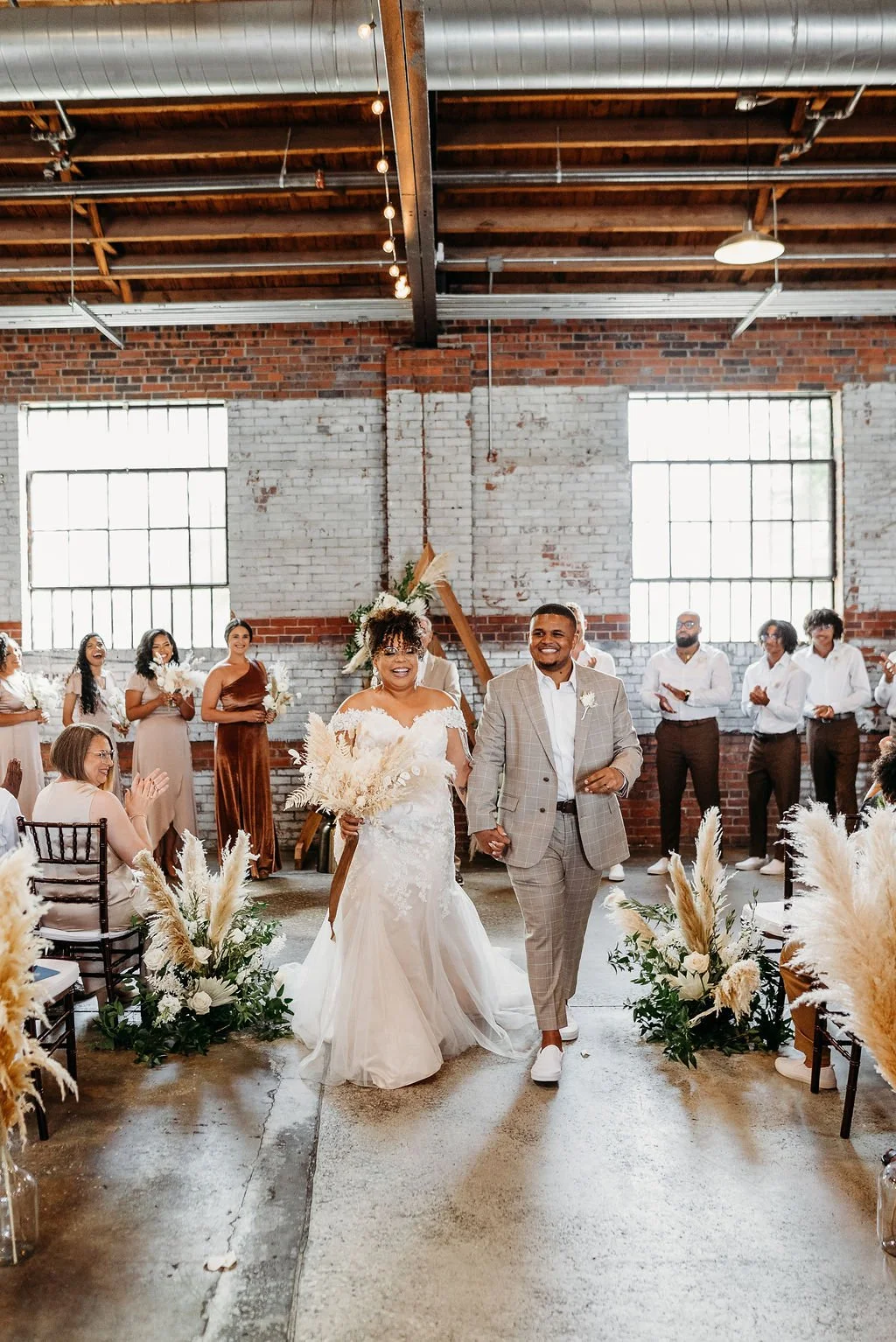 Industrial Chic wedding ceremony at Studio 215 in Fayetteville NC featuring exposed brick, string lights, a happy couple with the bride in a mermaid bridal gown, with floral pompous grass accents