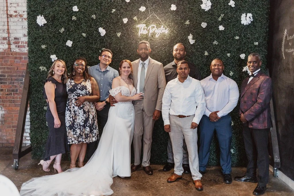 Group of people at a wedding, standing in front of a green leafy wall with white flowers and a glowing sign that reads 'The Poth.' The group includes bride and groom in the center, dressed in wedding attire, surrounded by friends or family.