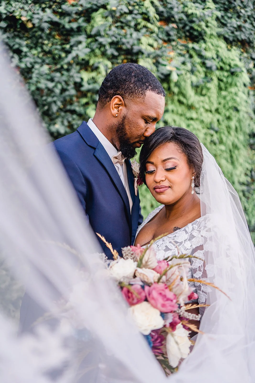 A bride and groom softly embrace outdoors, with the groom in a navy suit and the bride in a white lace gown and veil, holding a bouquet of pink and white flowers, in front of green foliage.