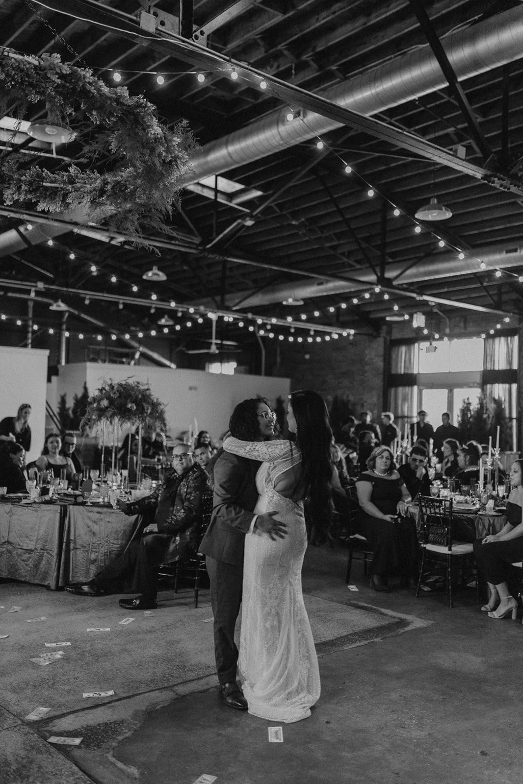 A black and white photo of a wedding reception. A couple is dancing in the center, surrounded by seated guests at decorated tables with candles and floral arrangements. The venue has an industrial style with string lights and high ceilings.
