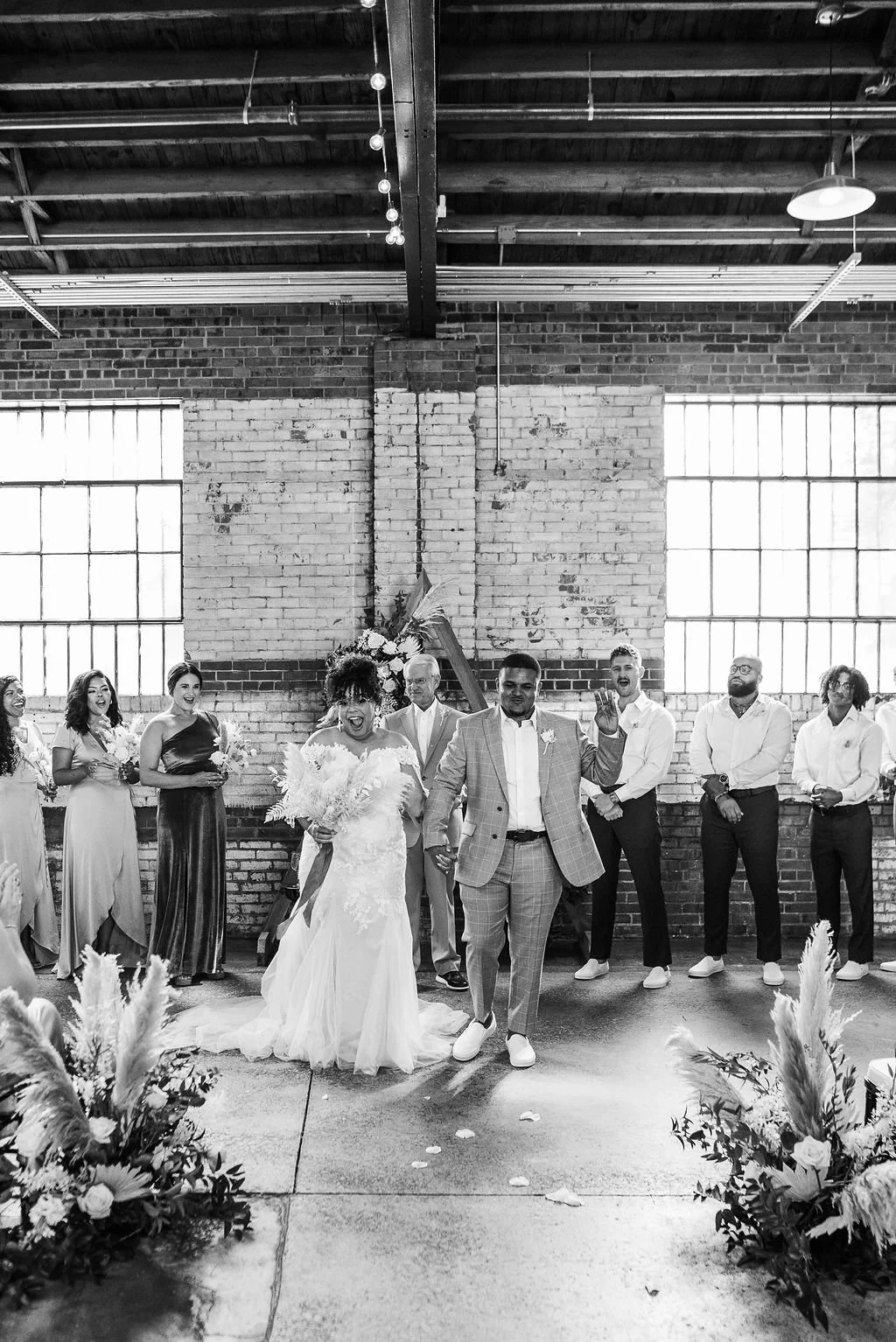 A wedding celebration with a bride and groom walking hand in hand, surrounded by bridesmaids and groomsmen in an industrial-style venue with large windows and exposed brick walls.