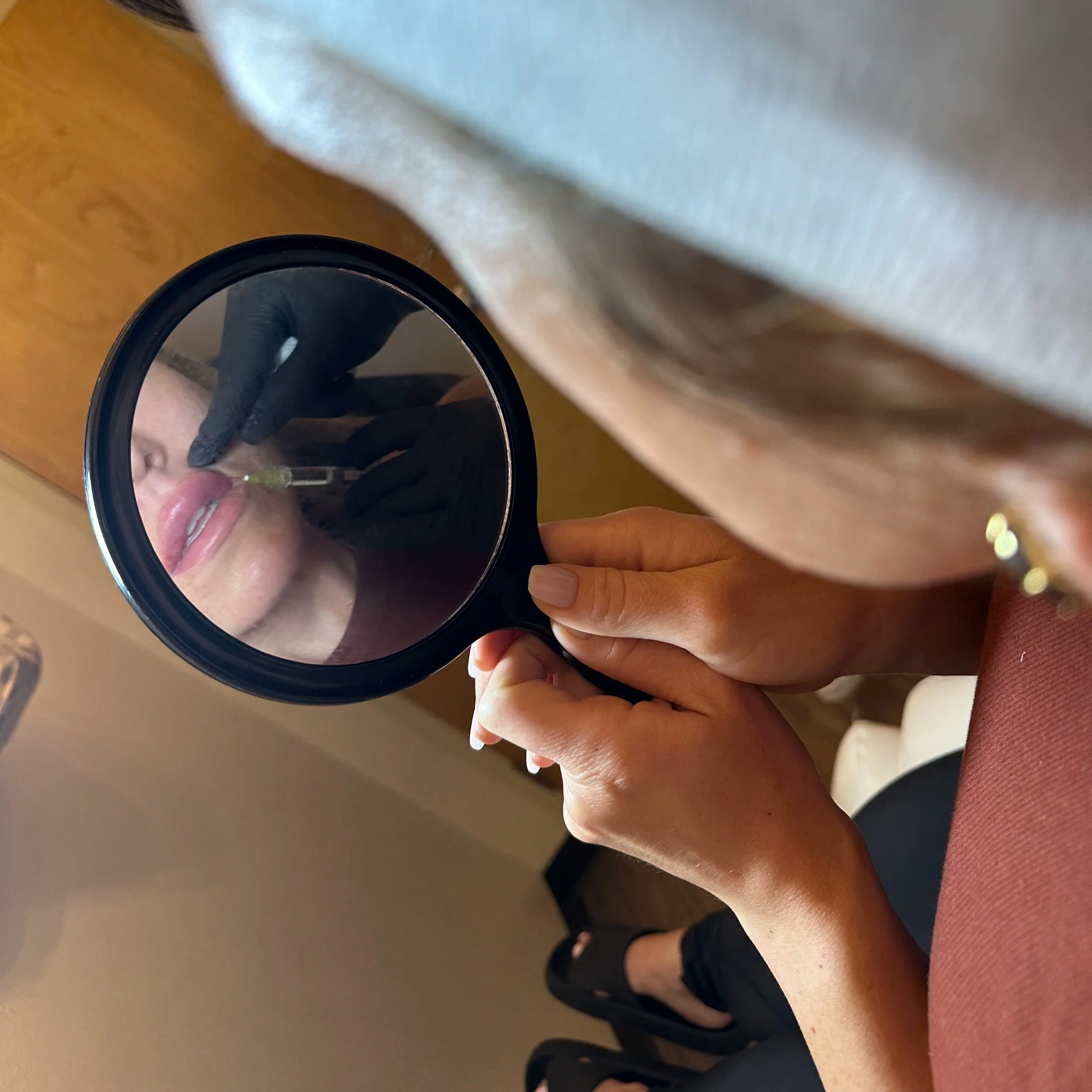 A woman holding a mirror captures her reflection as she is getting a lip injection from a medical professional wearing black gloves.