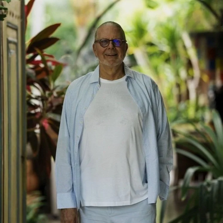 An elderly man with glasses and a beard, smiling, standing in a lush green garden, wearing a white t-shirt and a light blue button-up shirt.
