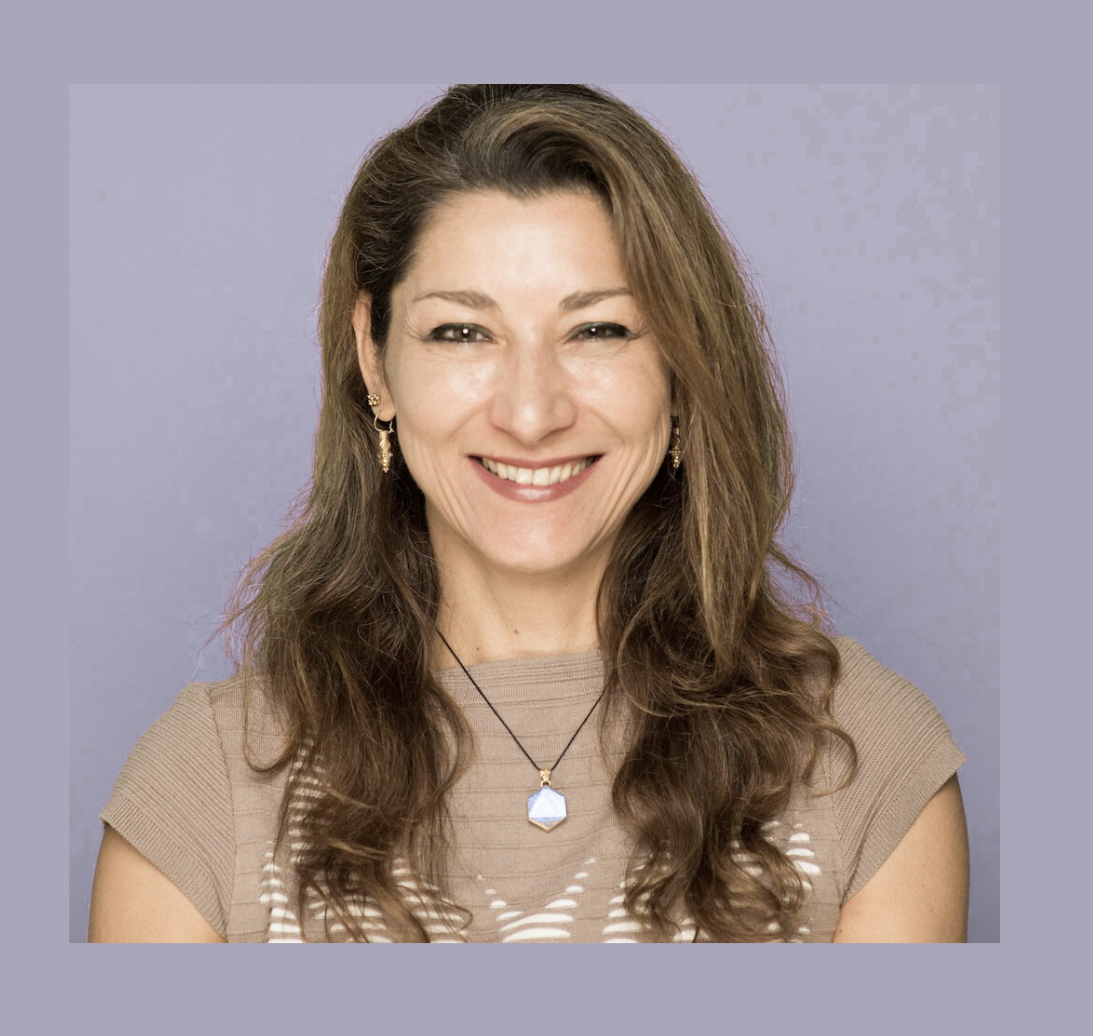 A woman with long, wavy brown hair, smiling, wearing a beige top, earrings, and a necklace, against a purple background.