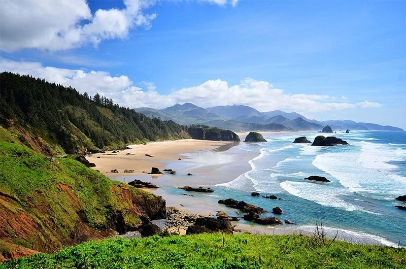 Scenic view of a sandy beach with rocks, green cliffs, and forested hills under a blue sky with clouds.