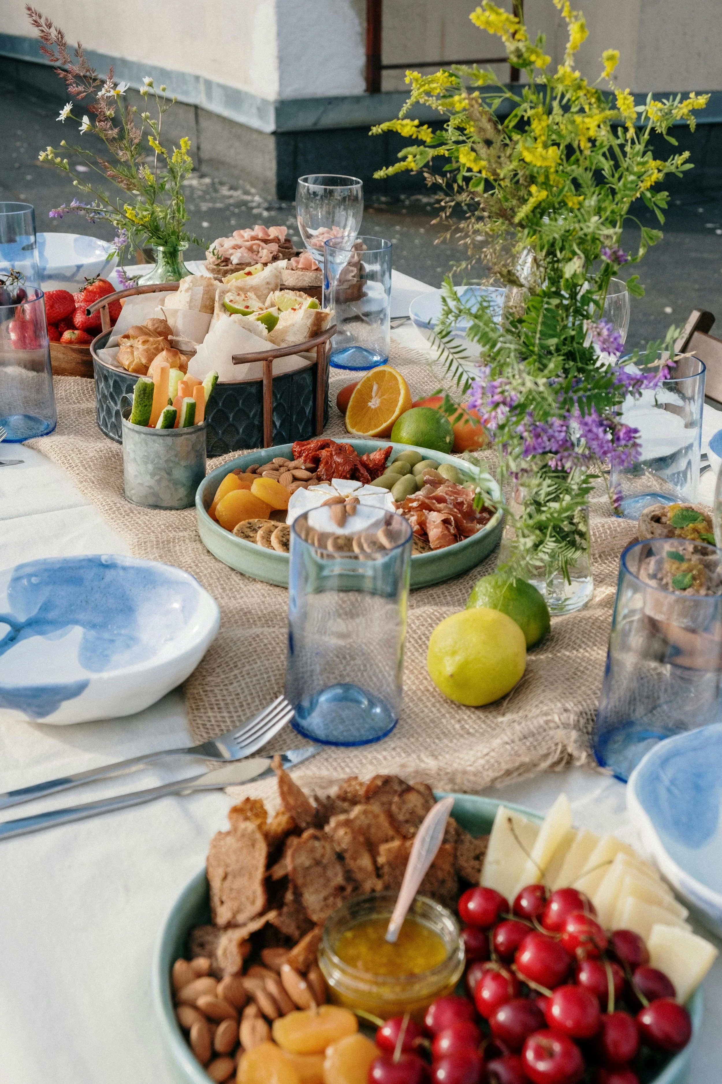 A colorful outdoor table set for a meal with various snacks, fruits, cheese, and flowers in vases.