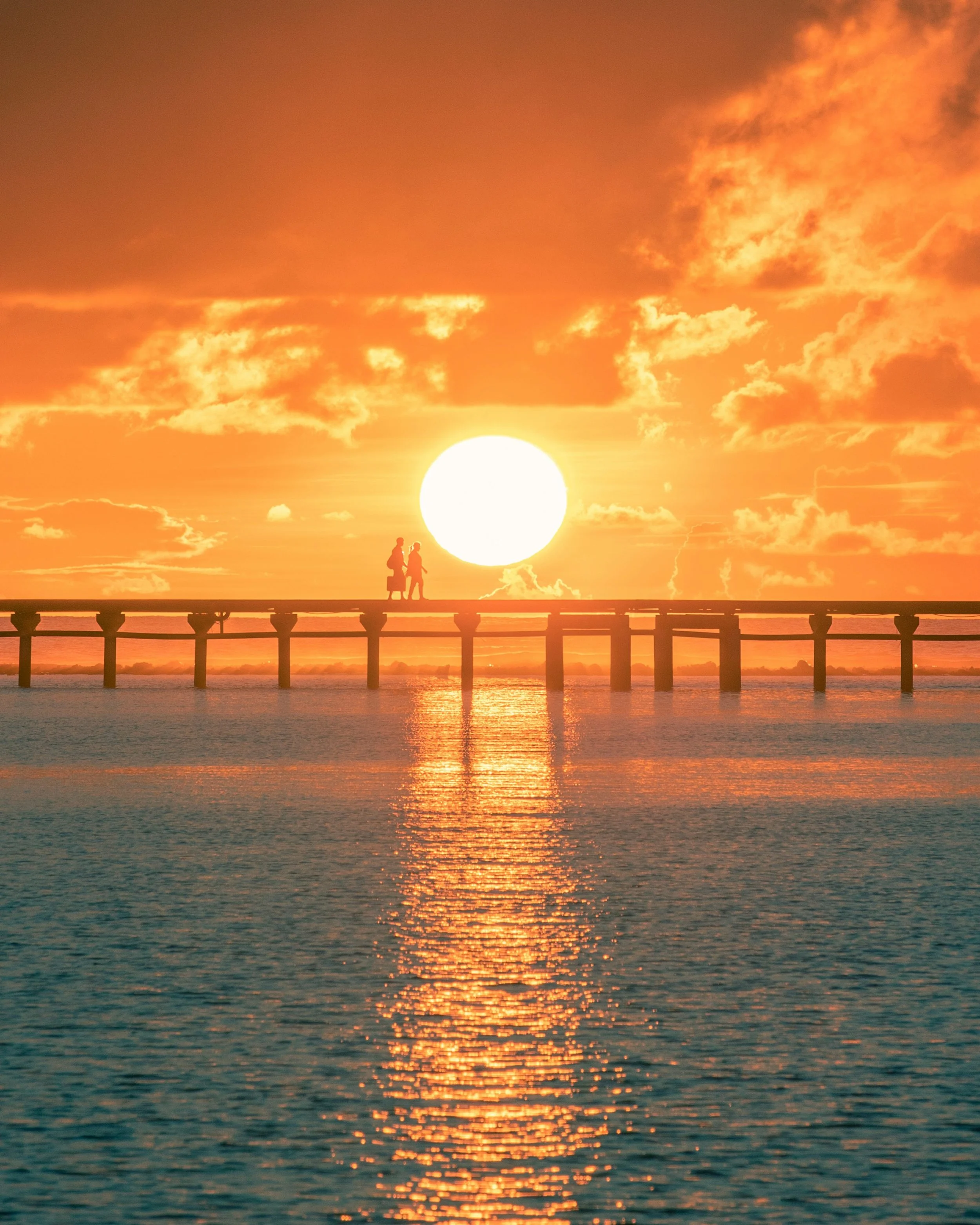Silhouette of two people walking on a pier at sunset over the ocean with orange and yellow sky and reflected water.