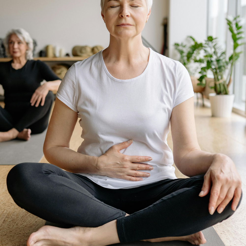 Older woman practicing meditation on a yoga mat, with hand on her stomach, eyes closed, in a bright room with plants.