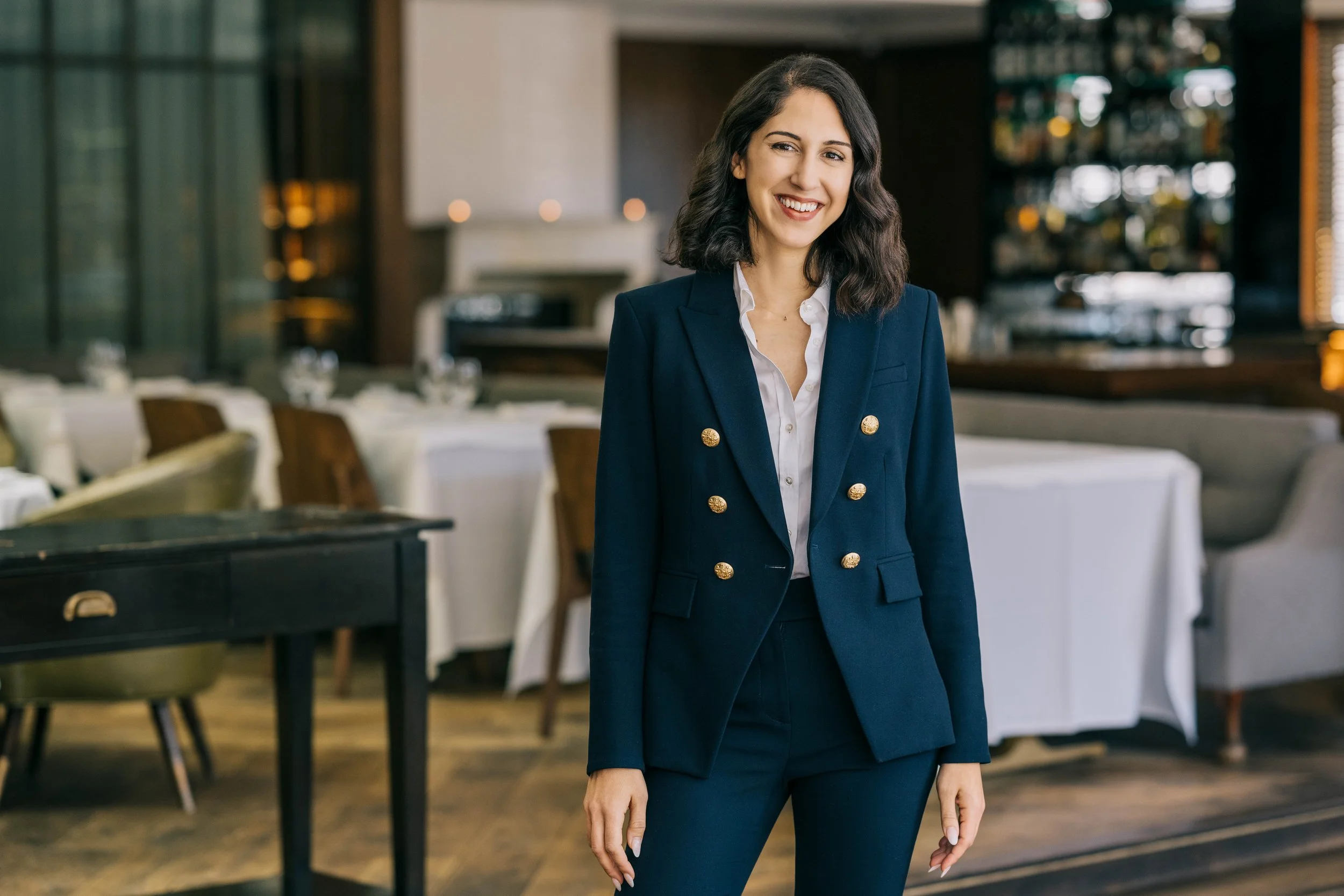 Photo of woman in a restaurant wearing a suit and smiling. Wood floors and white tablecloths.