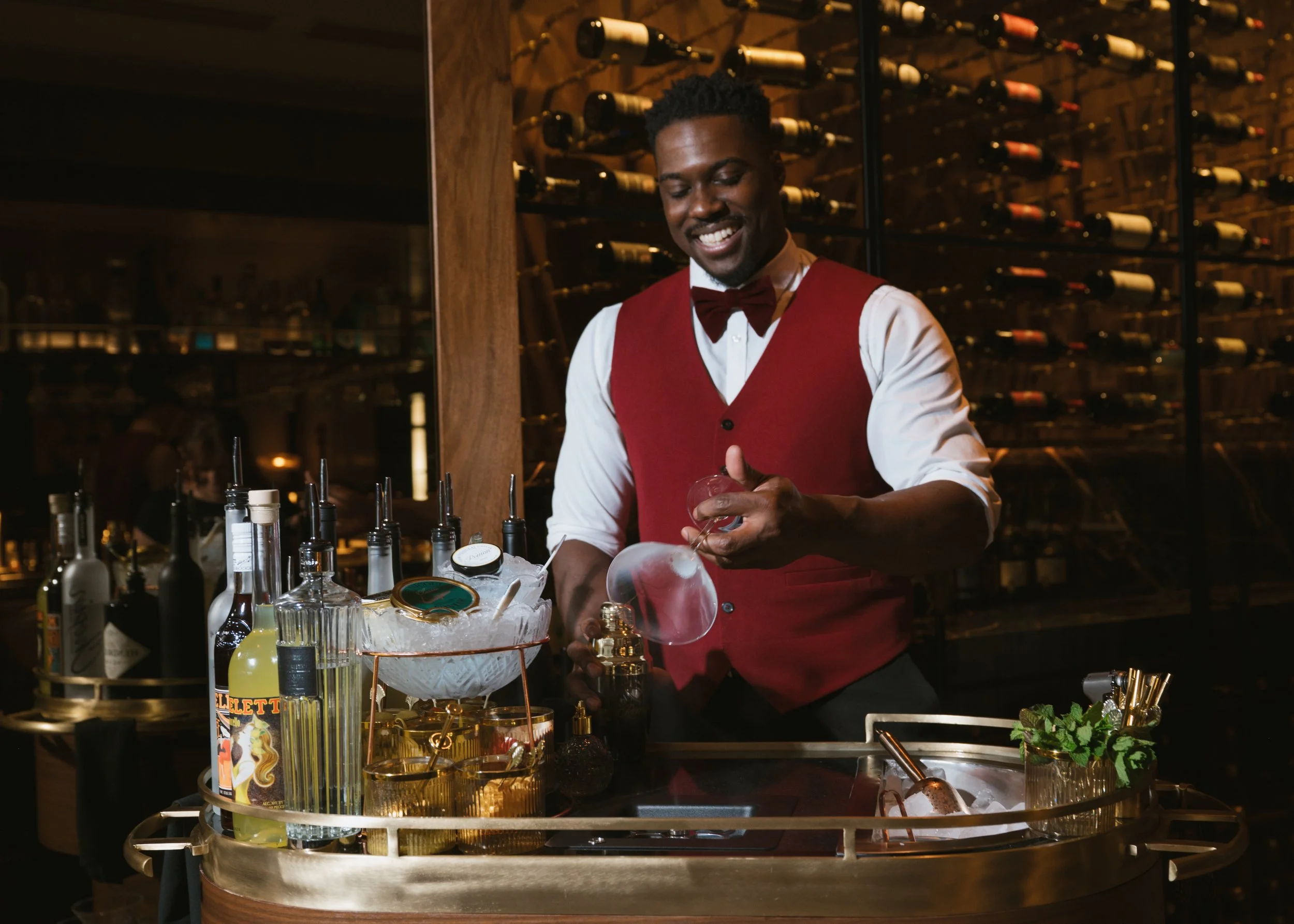 Man creating a martini at a martini cart inside a luxe restaurant with a wine cellar in the background