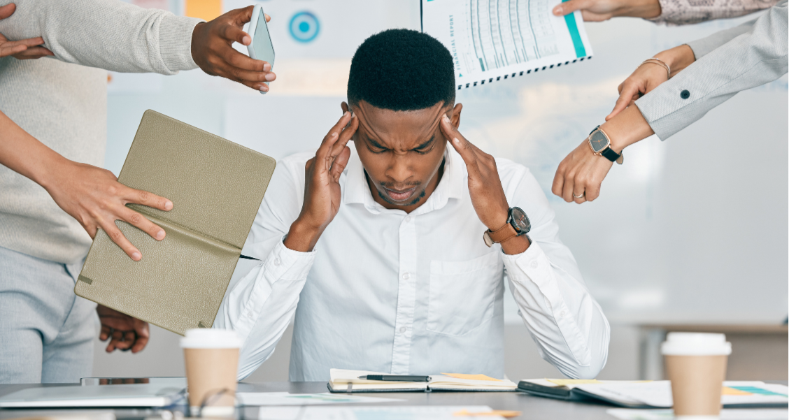 Un homme d'affaires stressé, entouré de plusieurs personnes lui montrant des documents et tenant un ordinateur portable et une tablette dans un environnement de bureau.