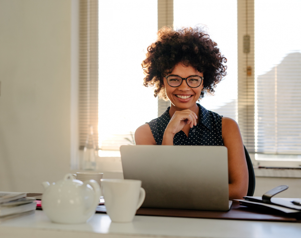 Femme souriante portant des lunettes, assise devant un ordinateur portable dans un bureau lumineux.