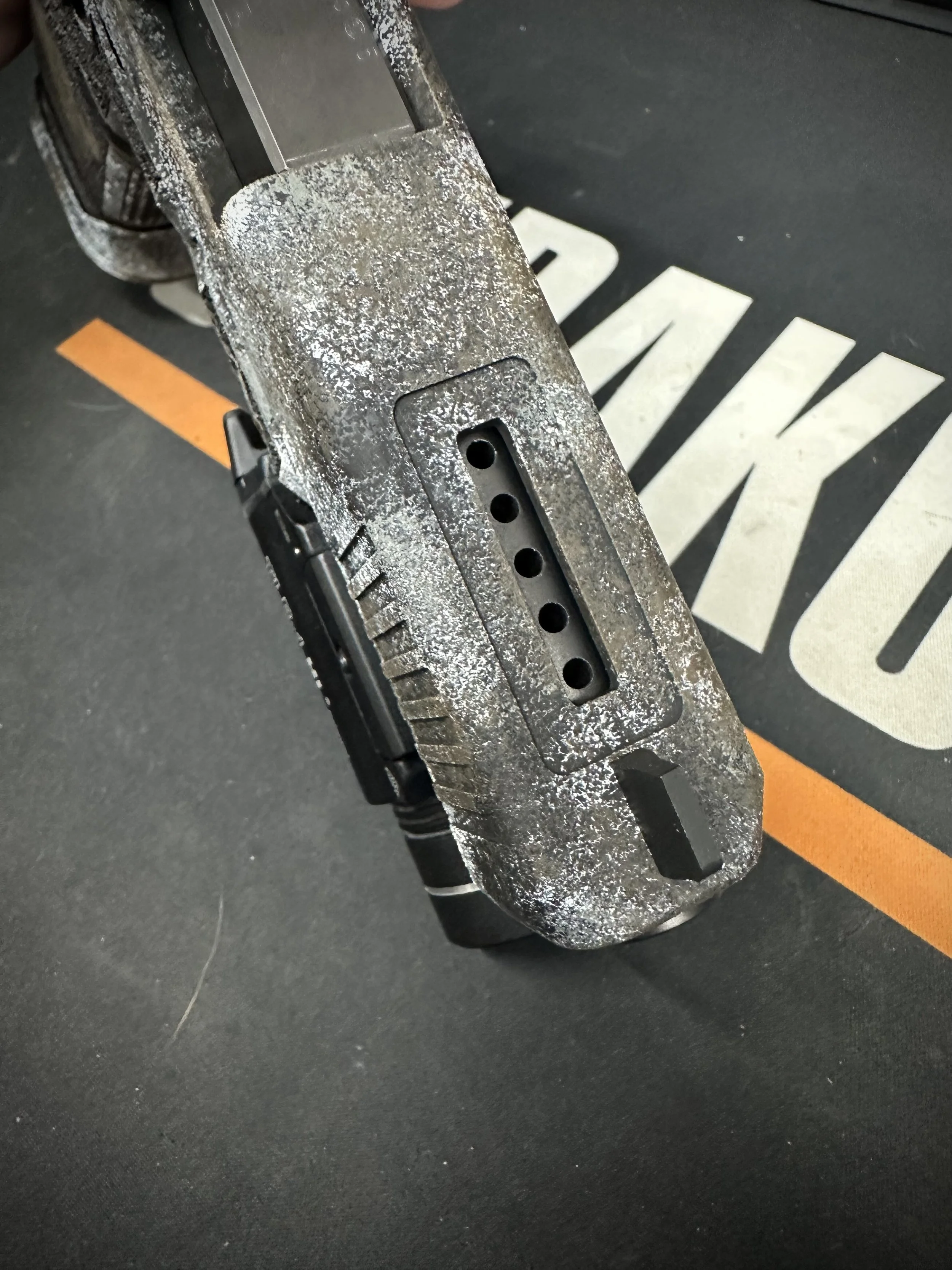 Close-up of a damaged skateboard with scratches and wear on the deck, placed on a black surface with orange and white markings.