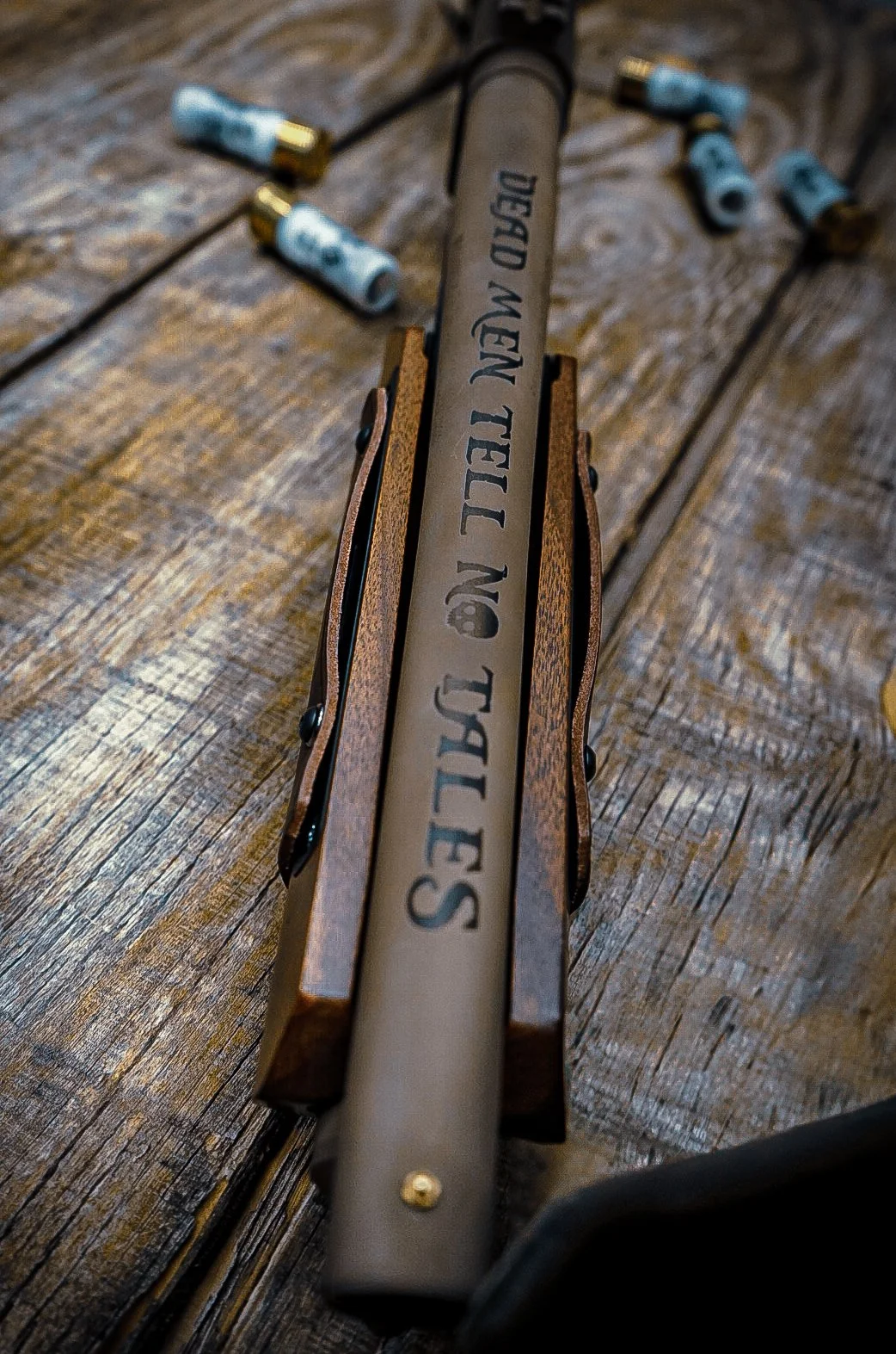 Close-up view of a firearm with the words 'DEAR MEN TELL NO TALLIES' engraved on the barrel, laid on a wooden surface with scattered small bullet casings around it.