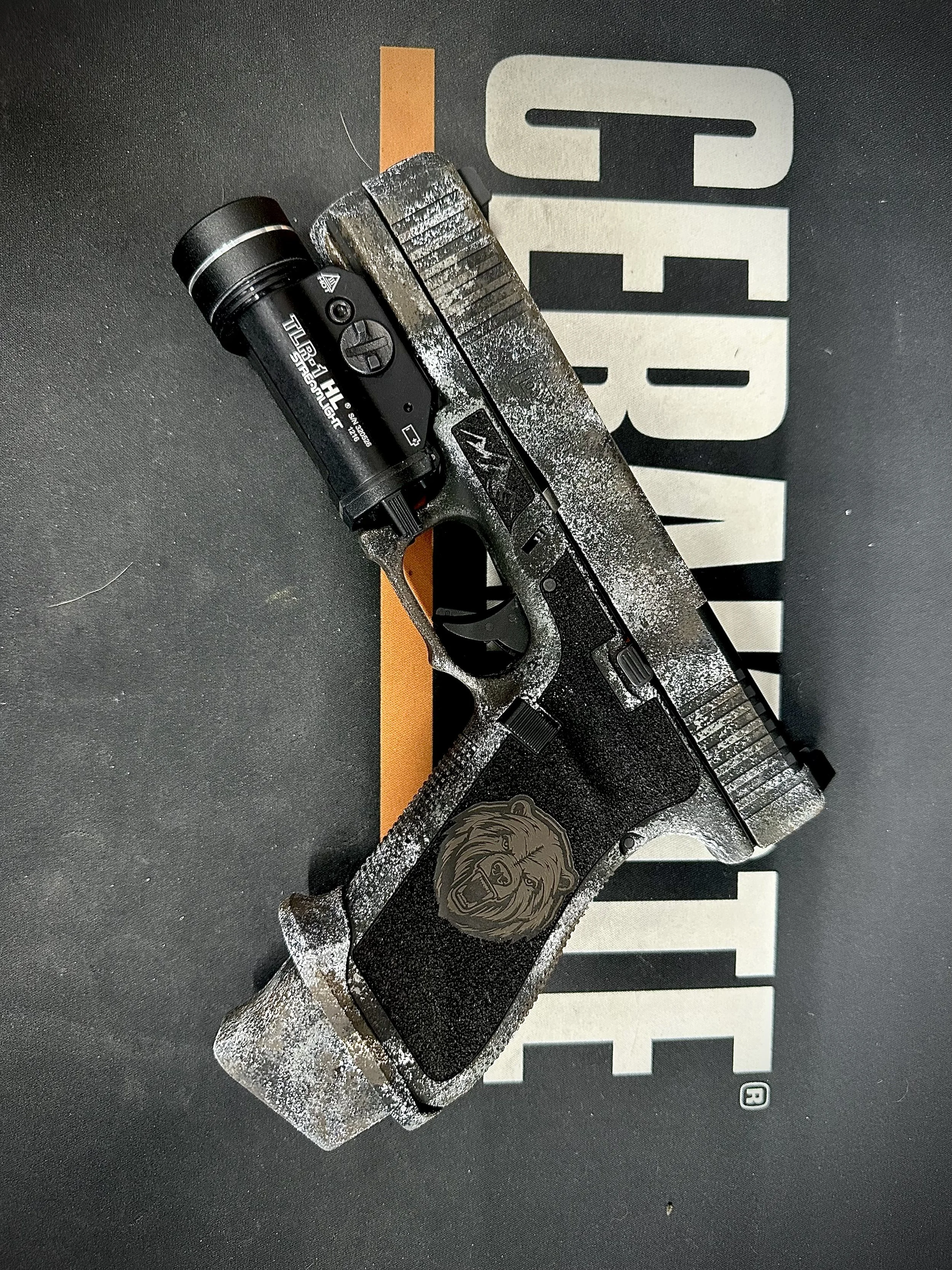 A black Glock pistol with a custom silver smoke finish, a lion emblem on the grip, and a flashlight mounted on the rail, resting on a dark surface with a partially visible large 