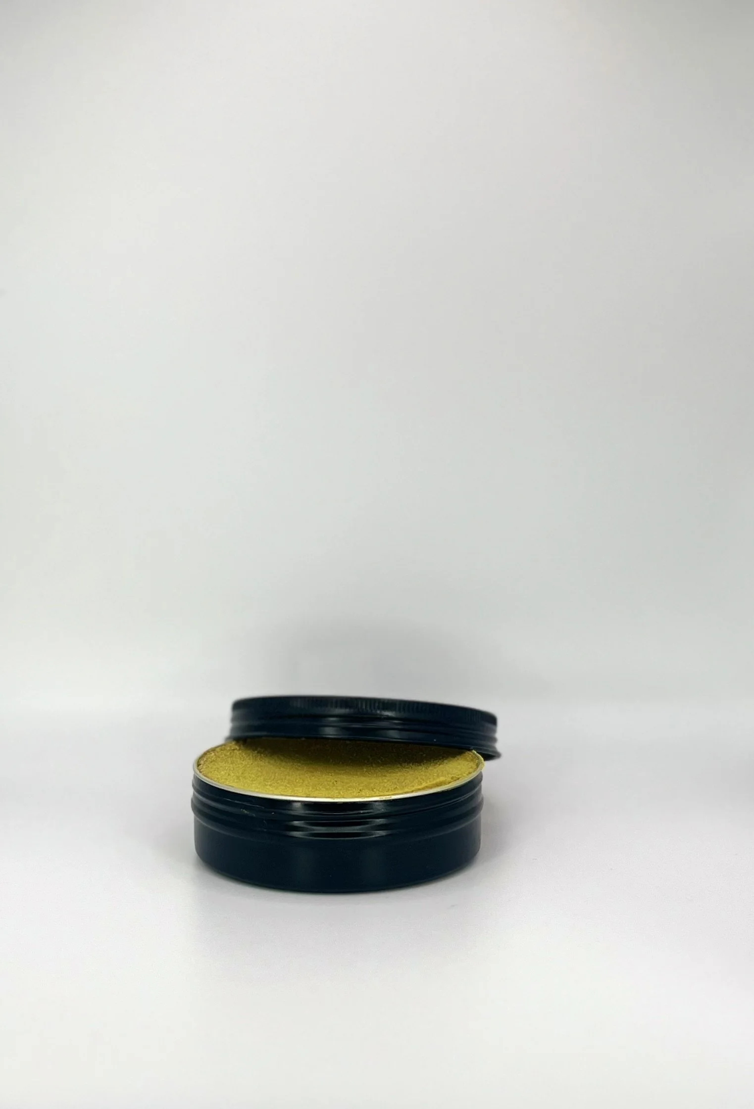 A black container with a lid, partially open, revealing a yellowish-green substance inside, placed on a white surface against a white background.