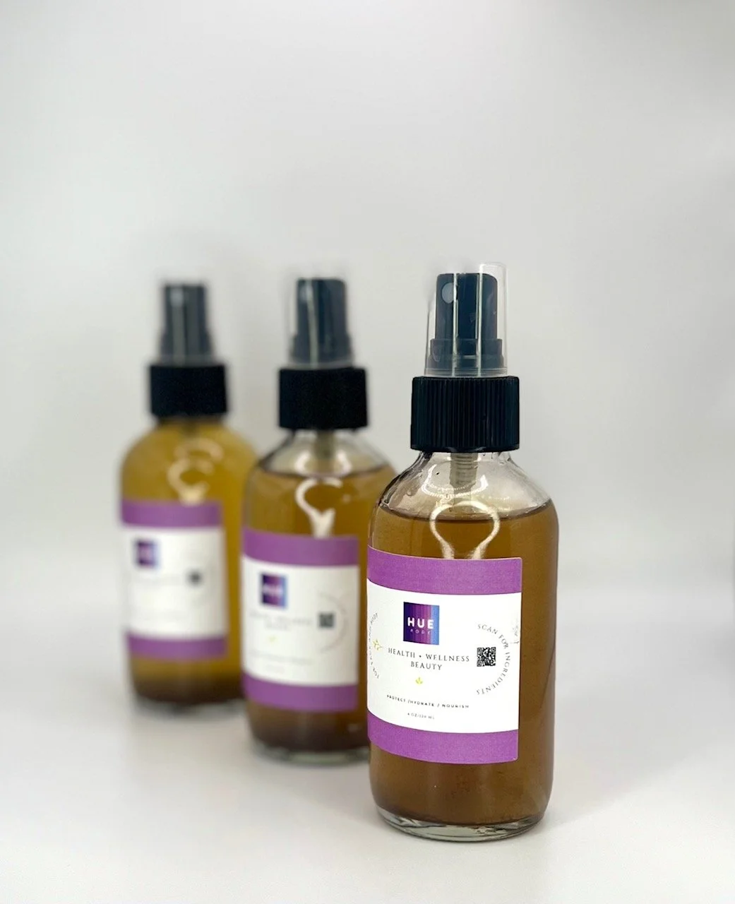 Three brown spray bottles with black caps and purple labels, arranged in a row on a white surface, one bottle in the foreground and two in the background.