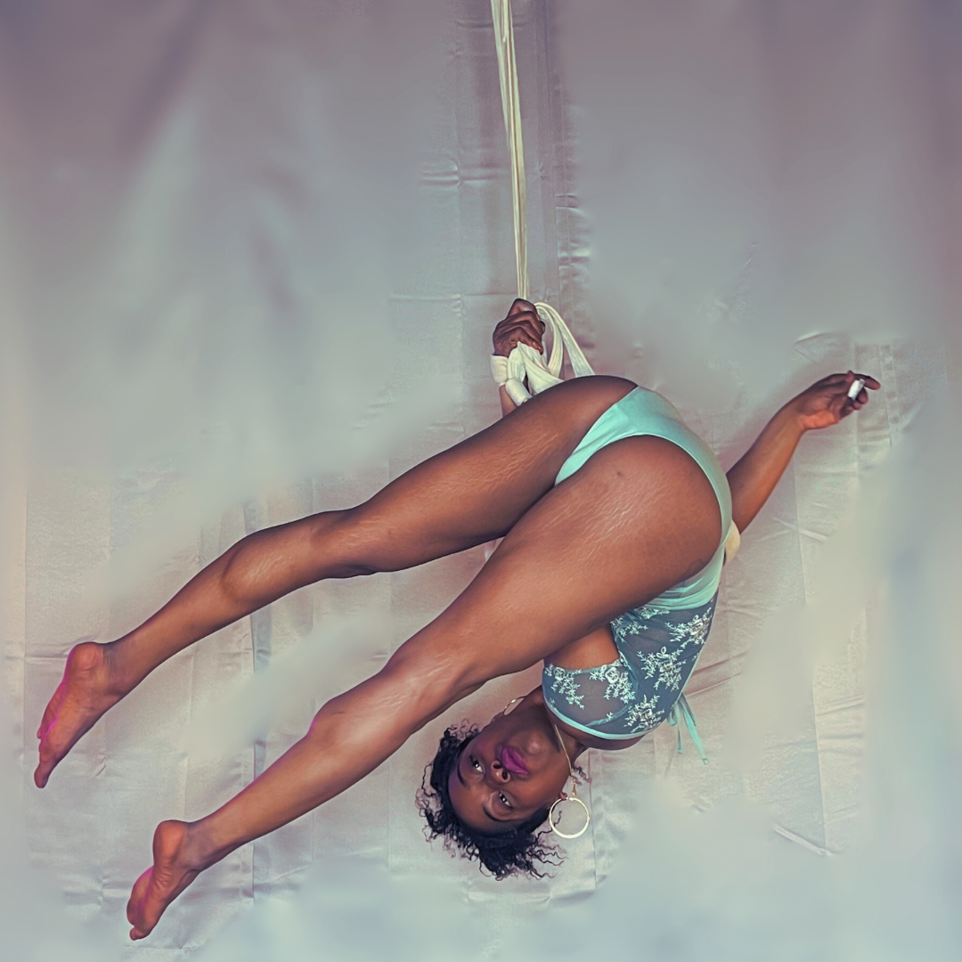 A woman performing aerial yoga against a neutral backdrop, hanging upside down with her legs spread apart and holding onto a yellow aerial silk.