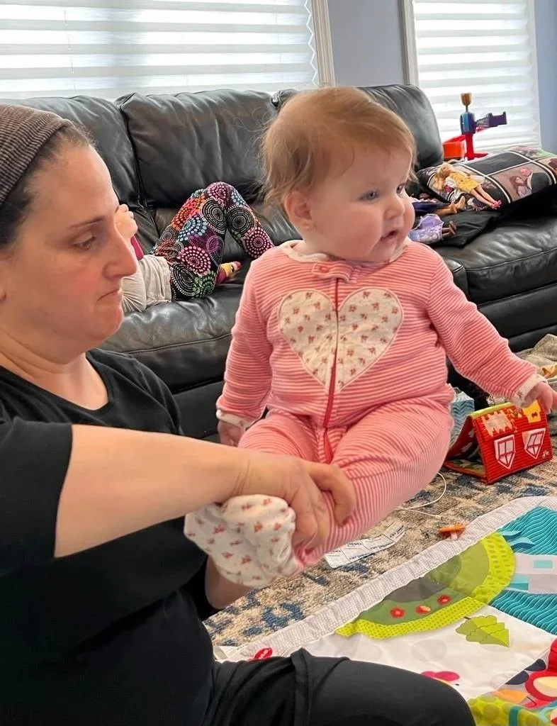 A woman holding a baby girl with reddish hair, wearing a pink striped outfit with a heart on the front, in a living room with toys and a couch in the background.