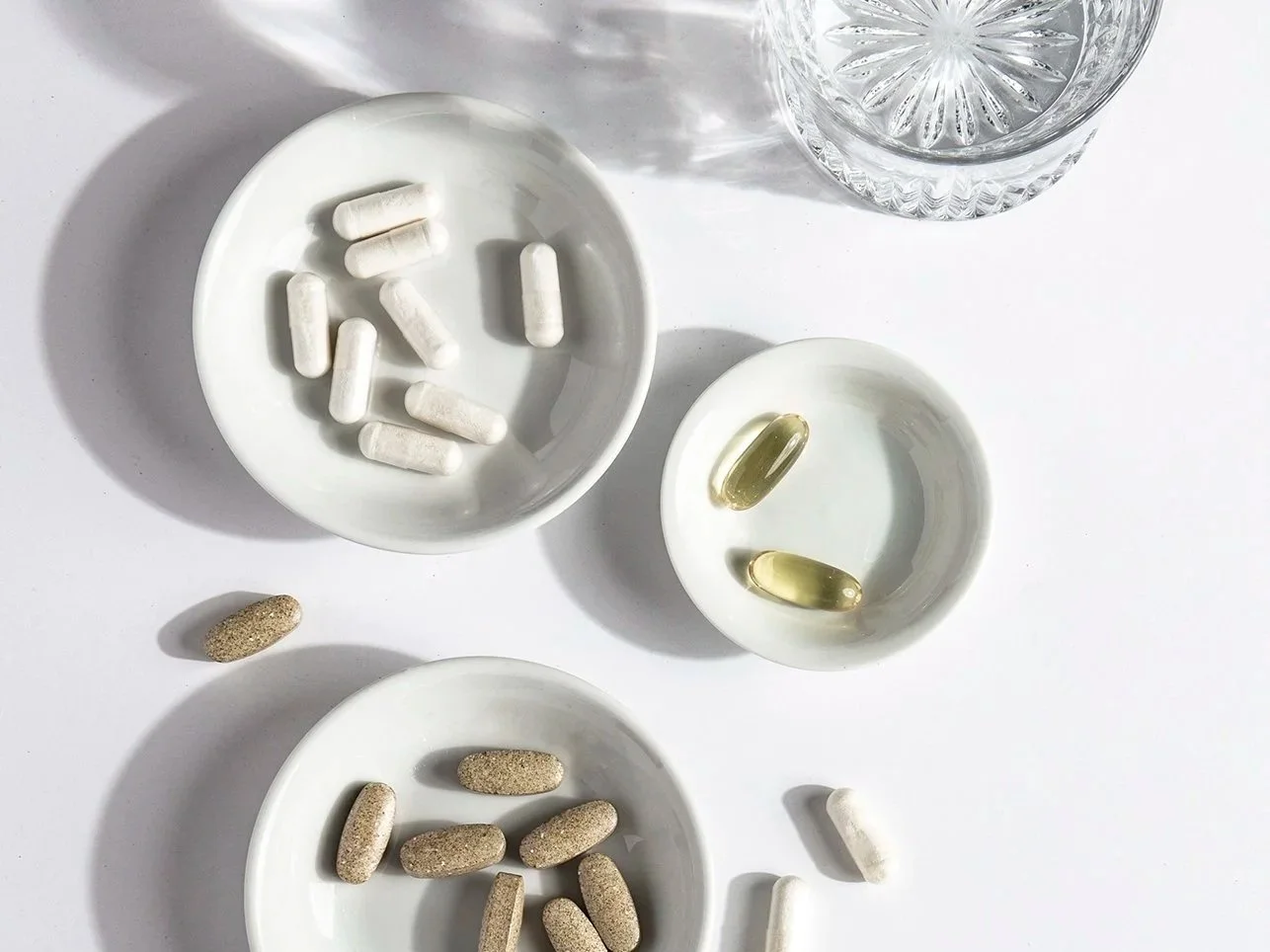 Various pills and capsules on white ceramic dishes, with a glass of water on a white surface.