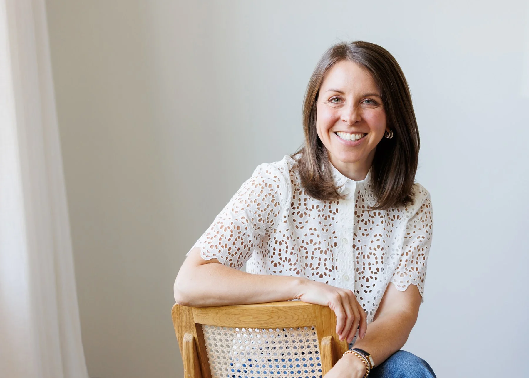 Smiling woman with brown hair sitting on a wooden chair in a white eyelet blouse against a plain background.