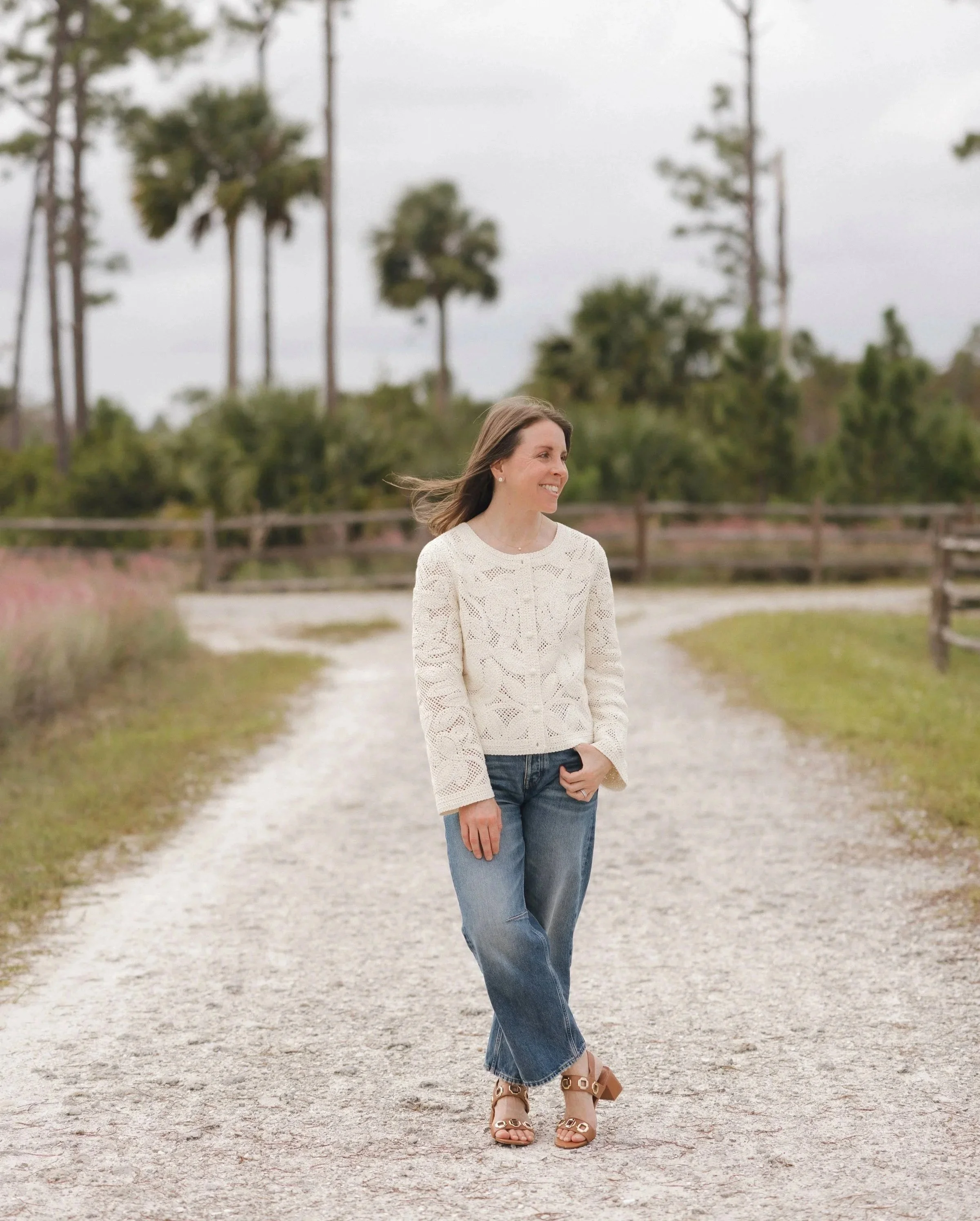 Woman walking on a dirt path in a park, wearing a white crochet sweater, jeans, and high heel sandals, with trees and fencing in the background.