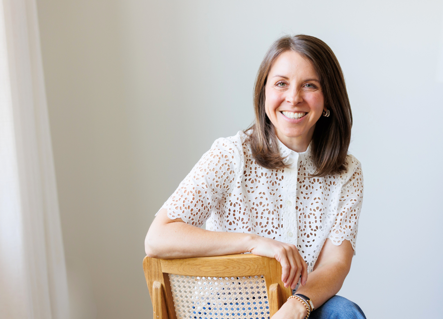 A woman smiling, sitting on a wooden chair with a cane back, wearing a white, short-sleeved, lace-patterned blouse, and jewelry, in a bright room with white walls.