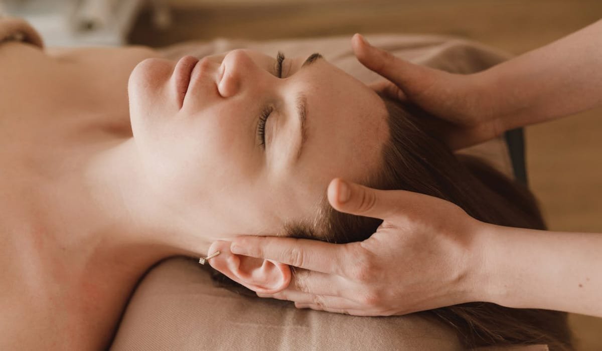 A woman receives a massage, lying on her back with eyes closed, while a therapist gently massages her head and neck.