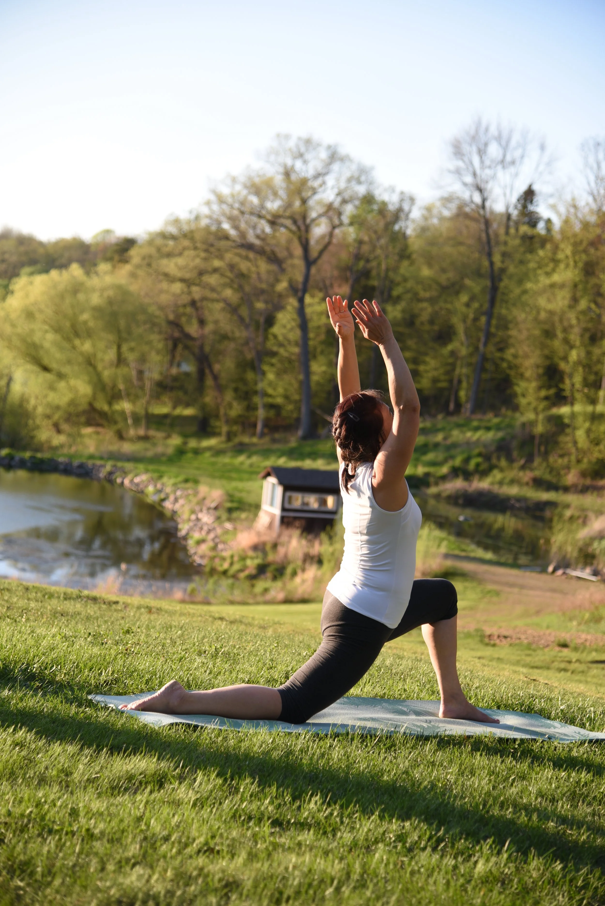 A woman practicing yoga outdoors on a grassy field near a lake, performing a lunge pose with arms raised upward during daylight.