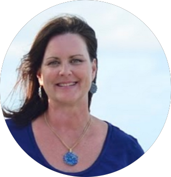 A woman smiling outdoors with her hair blowing in the wind, wearing a blue top and a large blue pendant necklace.