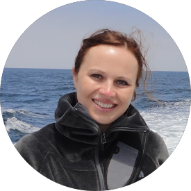 A woman with red hair smiling in front of the ocean, wearing a black jacket.
