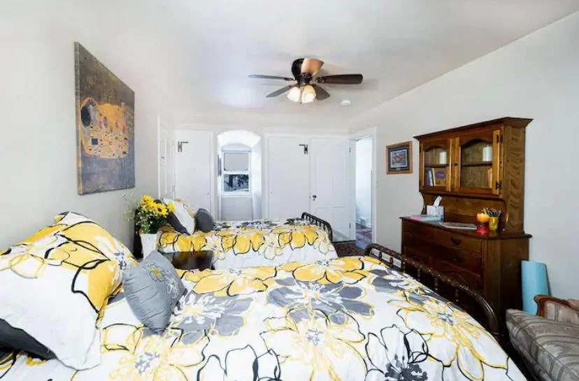 A bright bedroom with two beds featuring floral yellow, gray, and white bedding, a ceiling fan, a wooden dresser, a framed painting, and a window with an arched top.