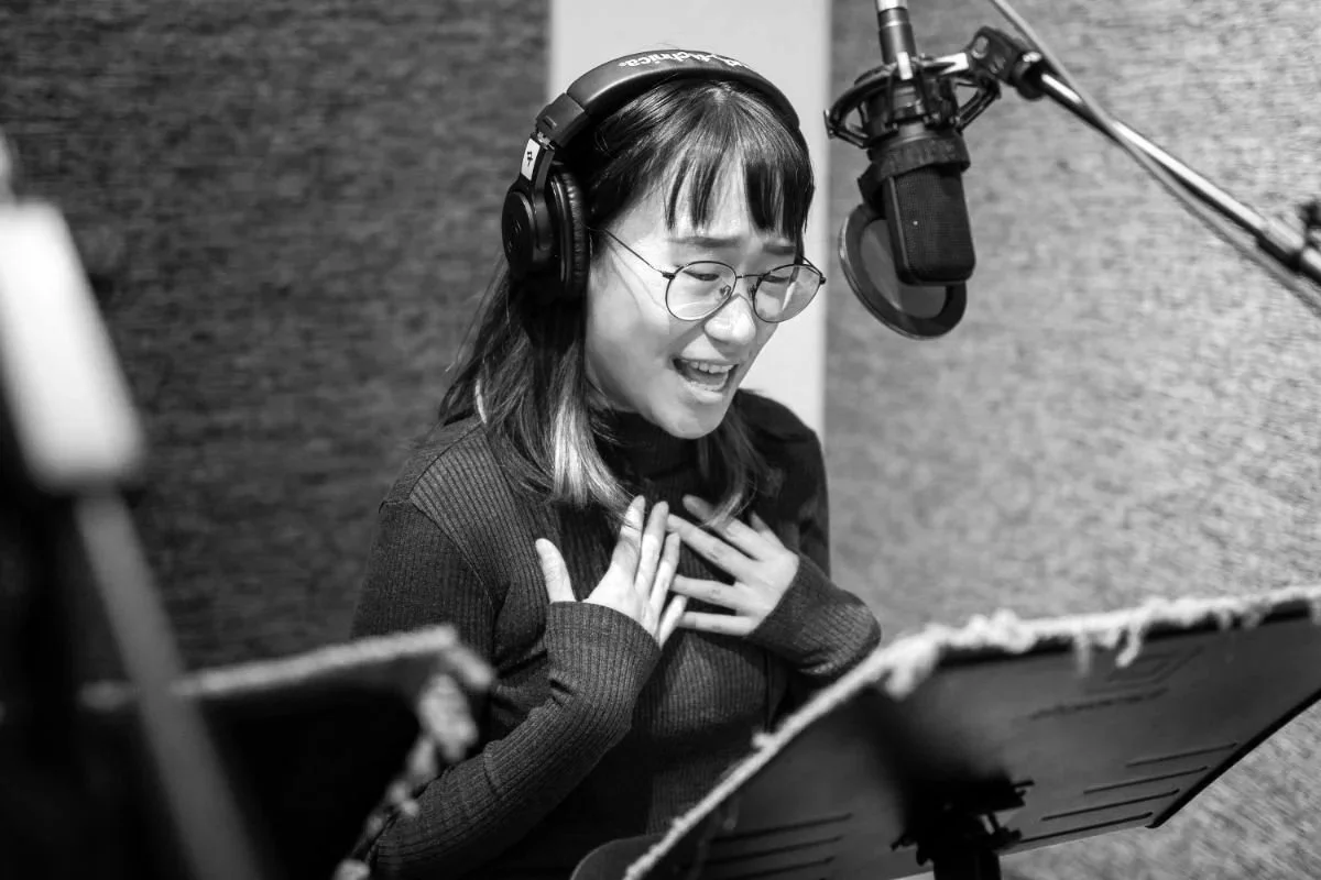 Young woman wearing headphones and performing expressively at a studio microphone.