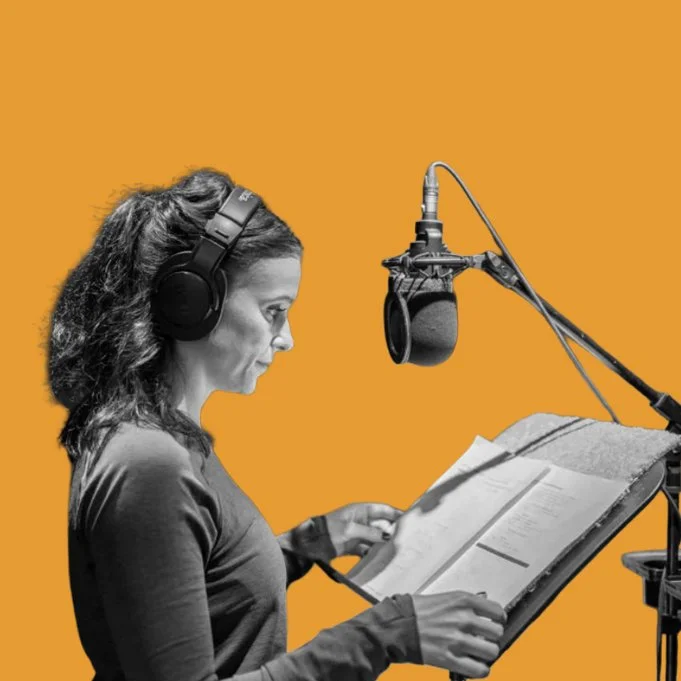Woman wearing headphones reading from a script beside a professional studio microphone.