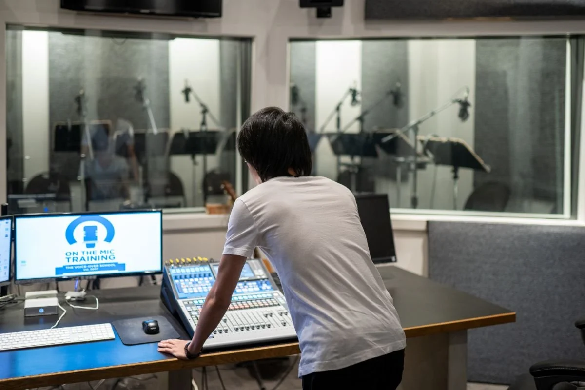 Audio engineer standing at a mixing console while looking into a professional recording studio through the control-room window.