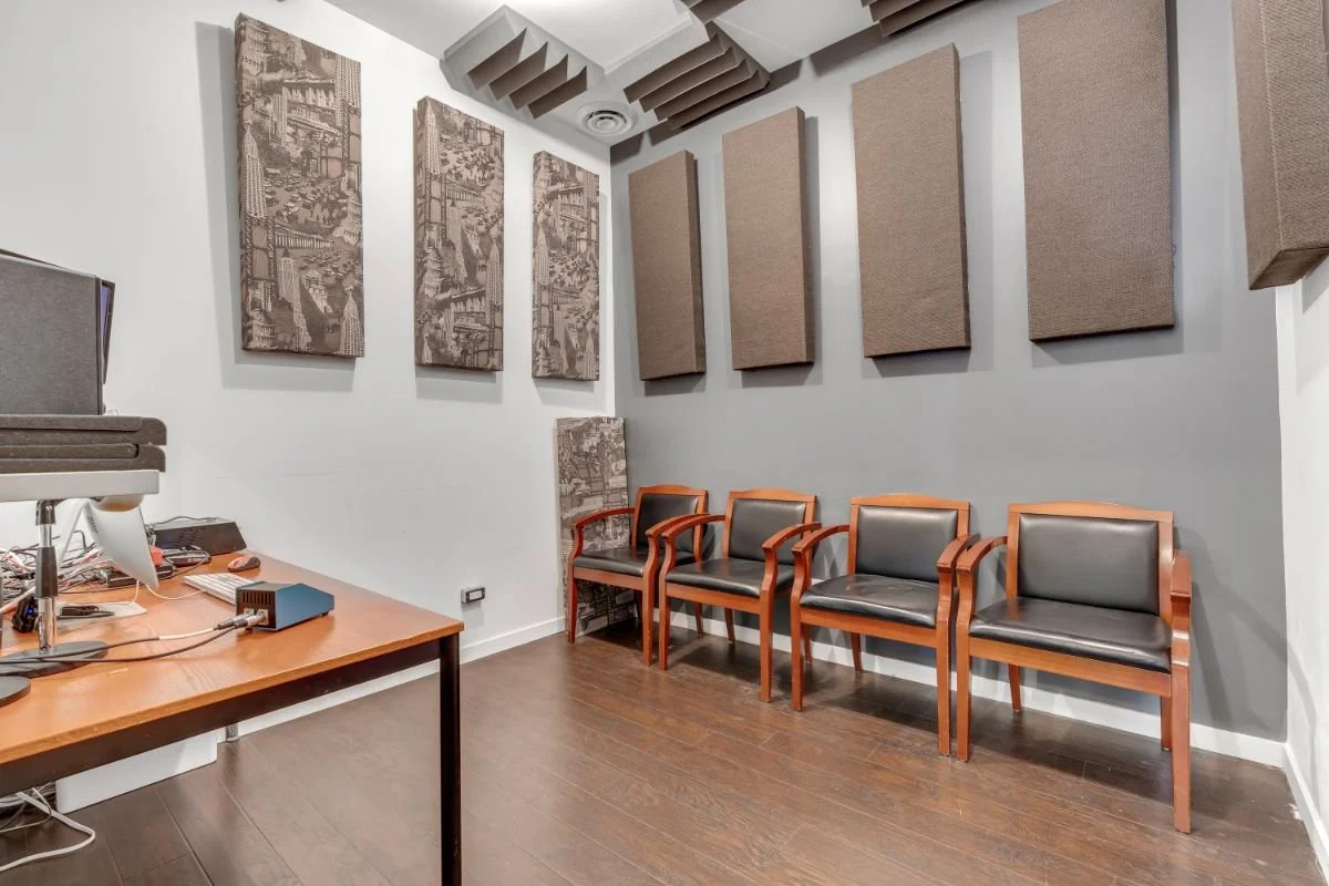 On The Mic Training studio with acoustic wall panels, three armchairs with wooden frames and black cushions, a wooden table with sound equipment, and a portion of another chair or stool in the corner.
