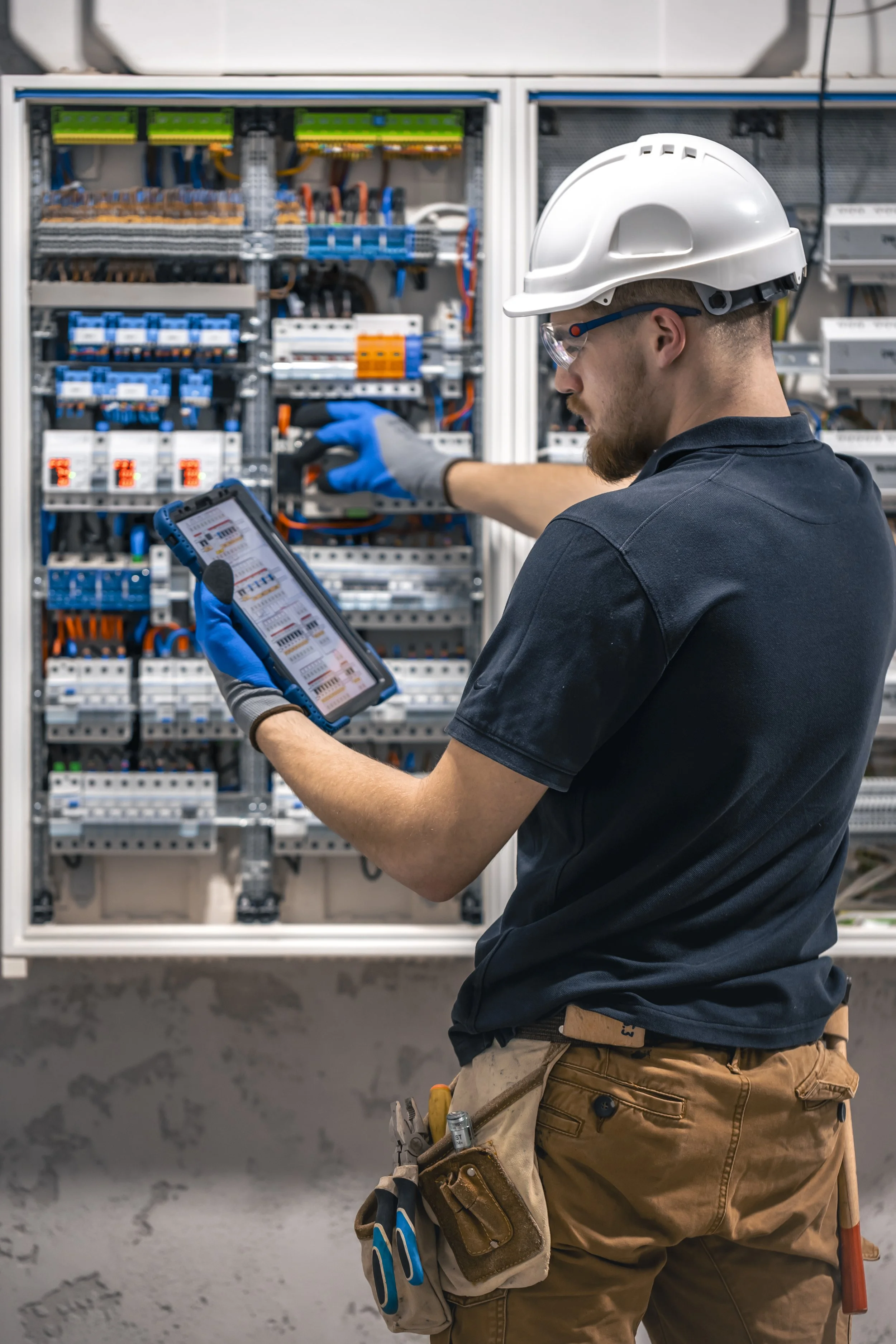 An electrician in a white hard hat and safety glasses inspects an open electrical panel while using a tablet.