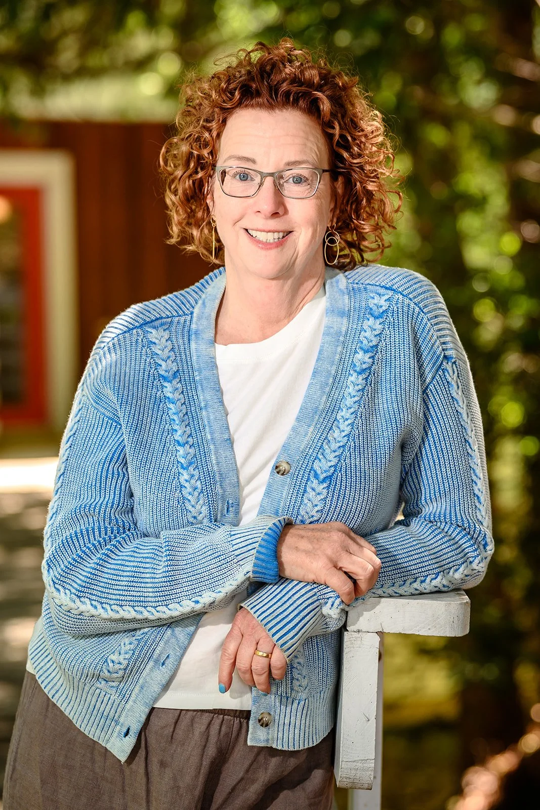 A woman with red curly hair and glasses wearing a blue sweater and leaning on a rail in the sunlight