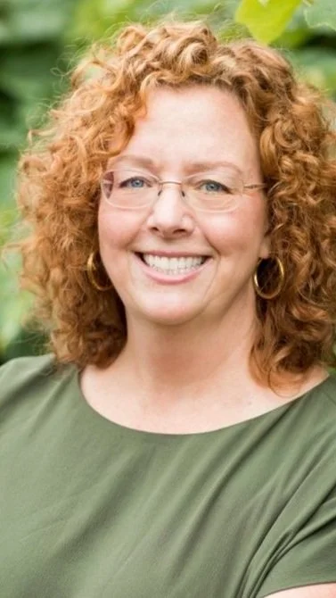 A woman with curly red hair wearing glasses and a green top, smiling outdoors among green plants.