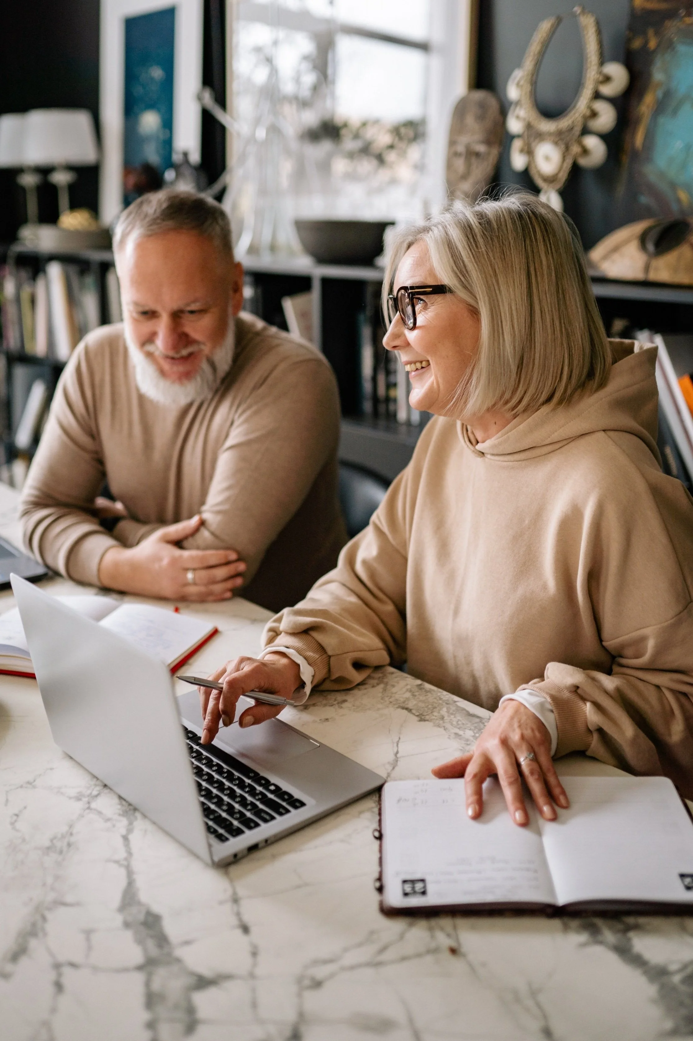 A woman with blonde hair and glasses smiling and pointing at a laptop screen, with a man with a gray beard smiling beside her, sitting at a table with notebooks. In a cozy room with bookshelves and decorative items in the background.