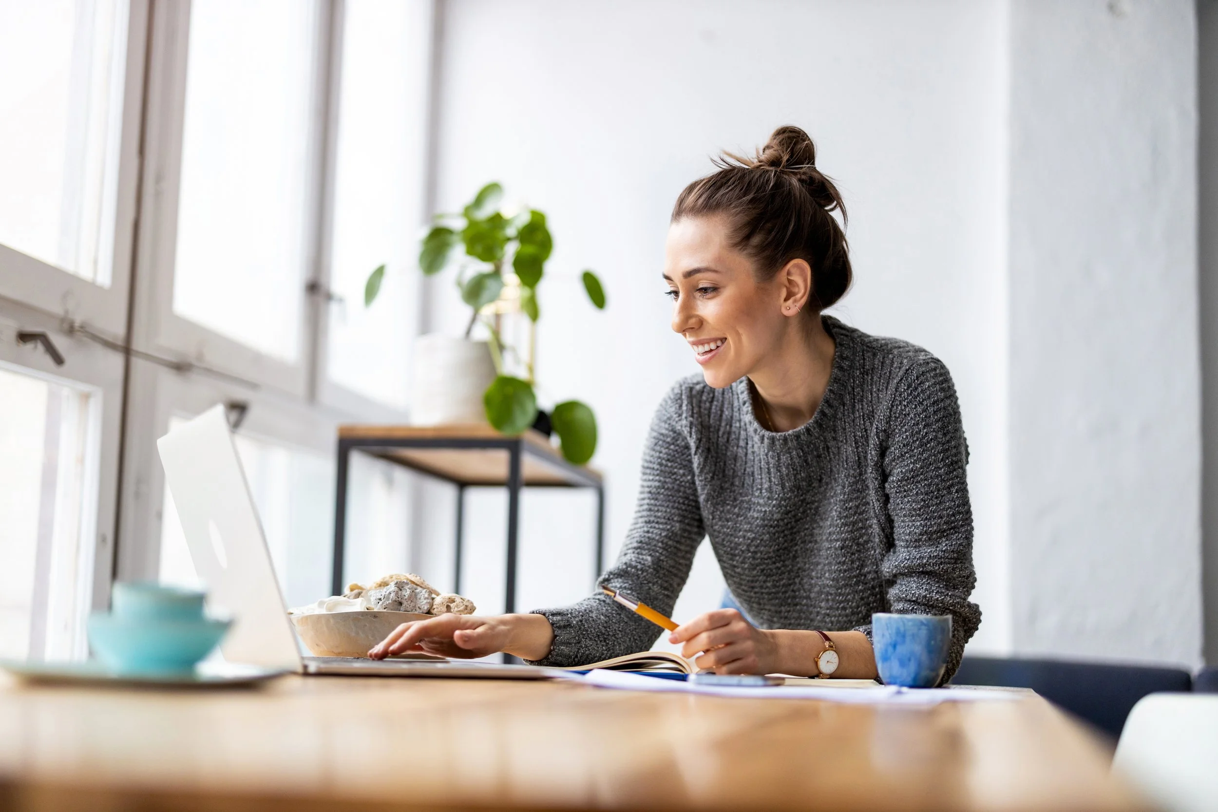 A woman sitting at a wooden table, working with a laptop, a pen in her hand and a smile on her face.