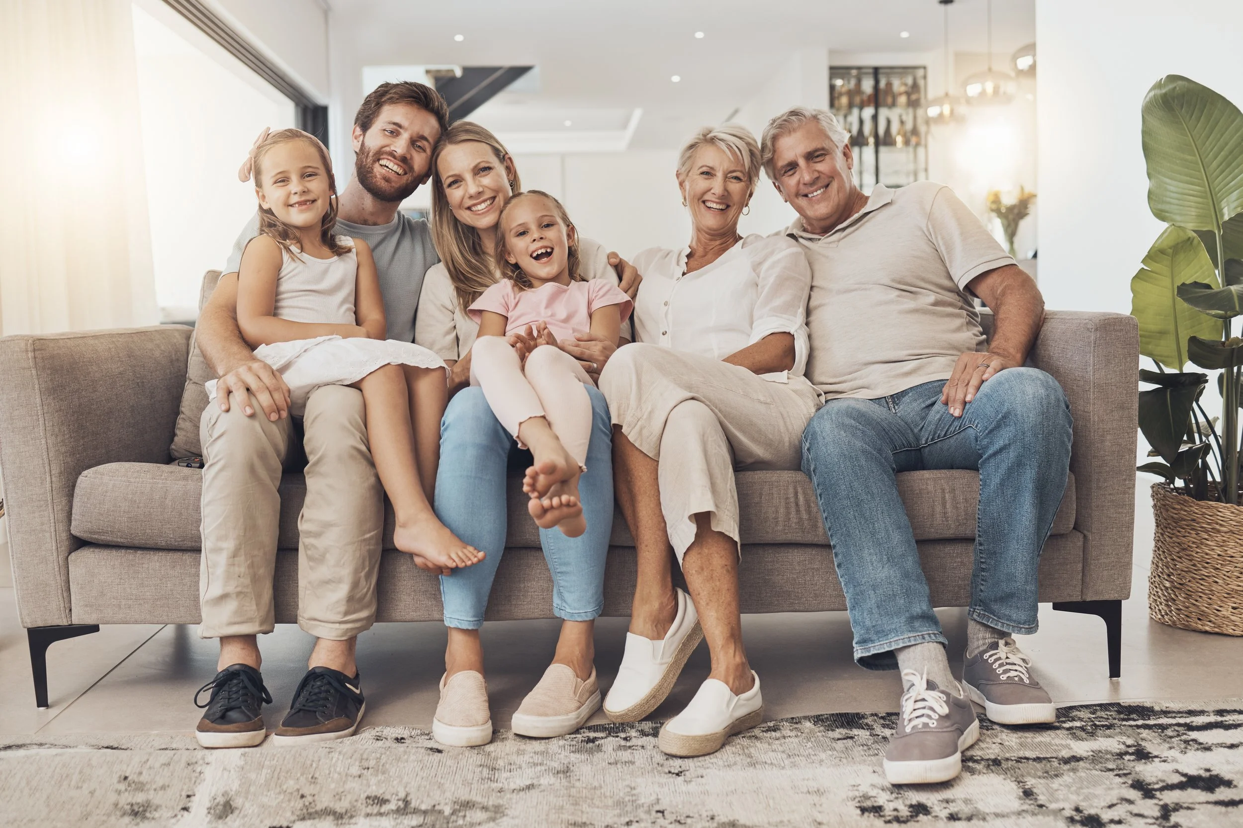 Family of six sitting on a beige sofa, smiling and enjoying each other's company in a bright living room.