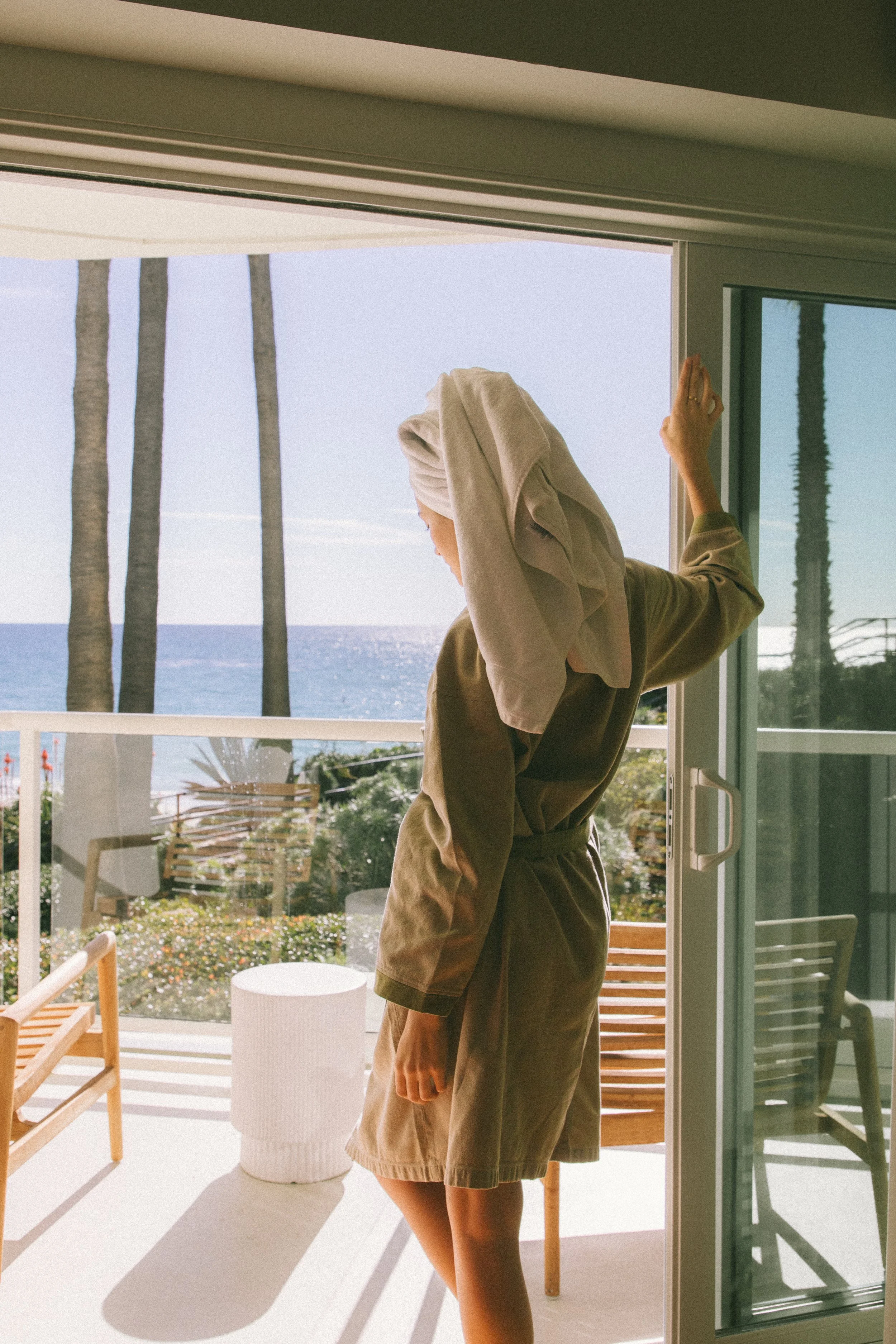 Person standing on a balcony overlooking the ocean, with palm trees in the background, seen from inside a room through a sliding door.