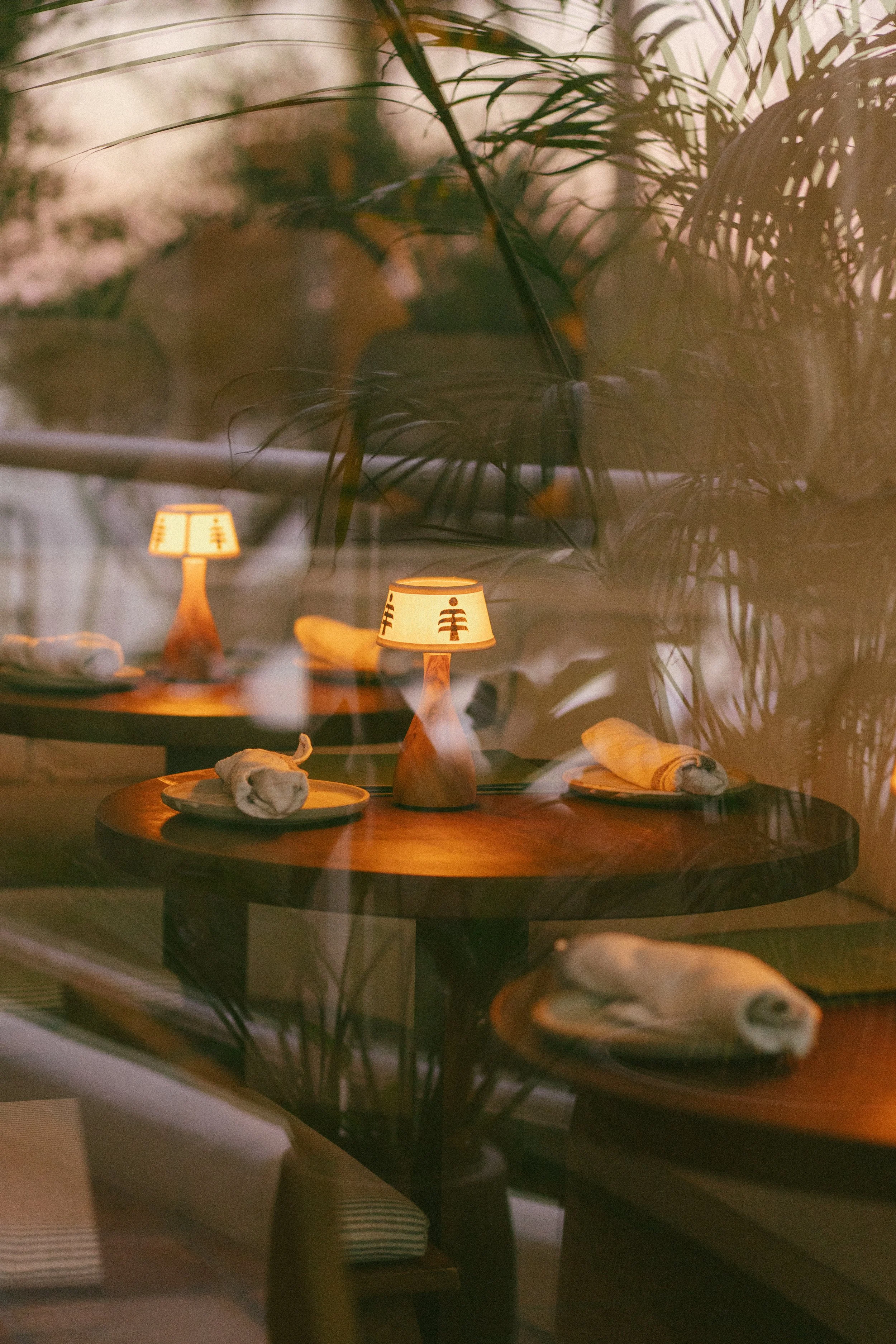 A cozy restaurant table setting with a lit small lamp, place settings with rolled napkins, and a view through the window showing reflections of the lamp and lush green plants.