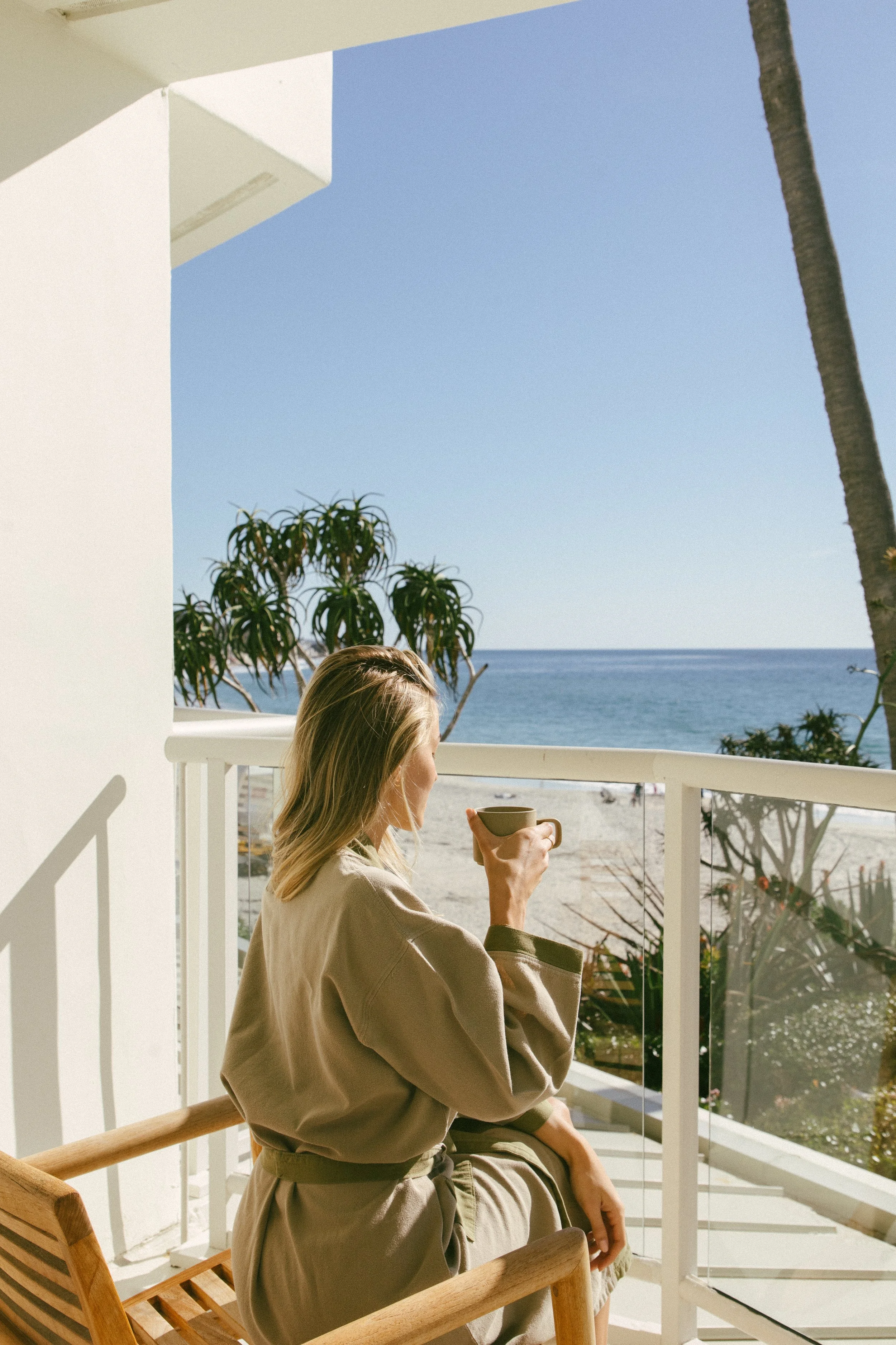 Woman sitting on a balcony overlooking the beach and ocean, holding a mug, with clear blue sky and palm trees in the background.