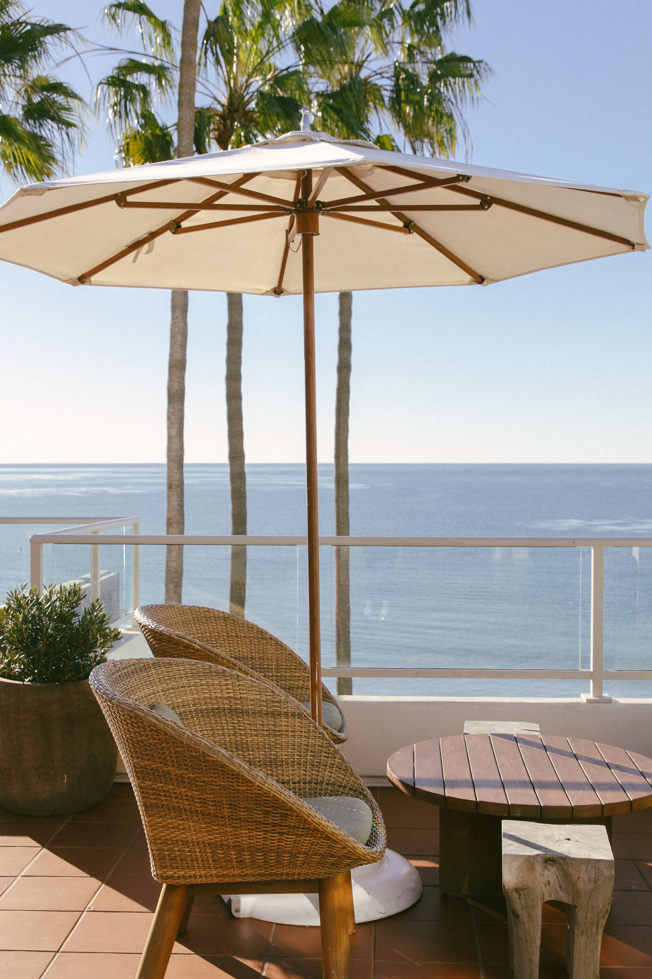 Beachside terrace with two wicker chairs, a small round table, large white patio umbrella, potted green plant, and ocean view with tall palm trees