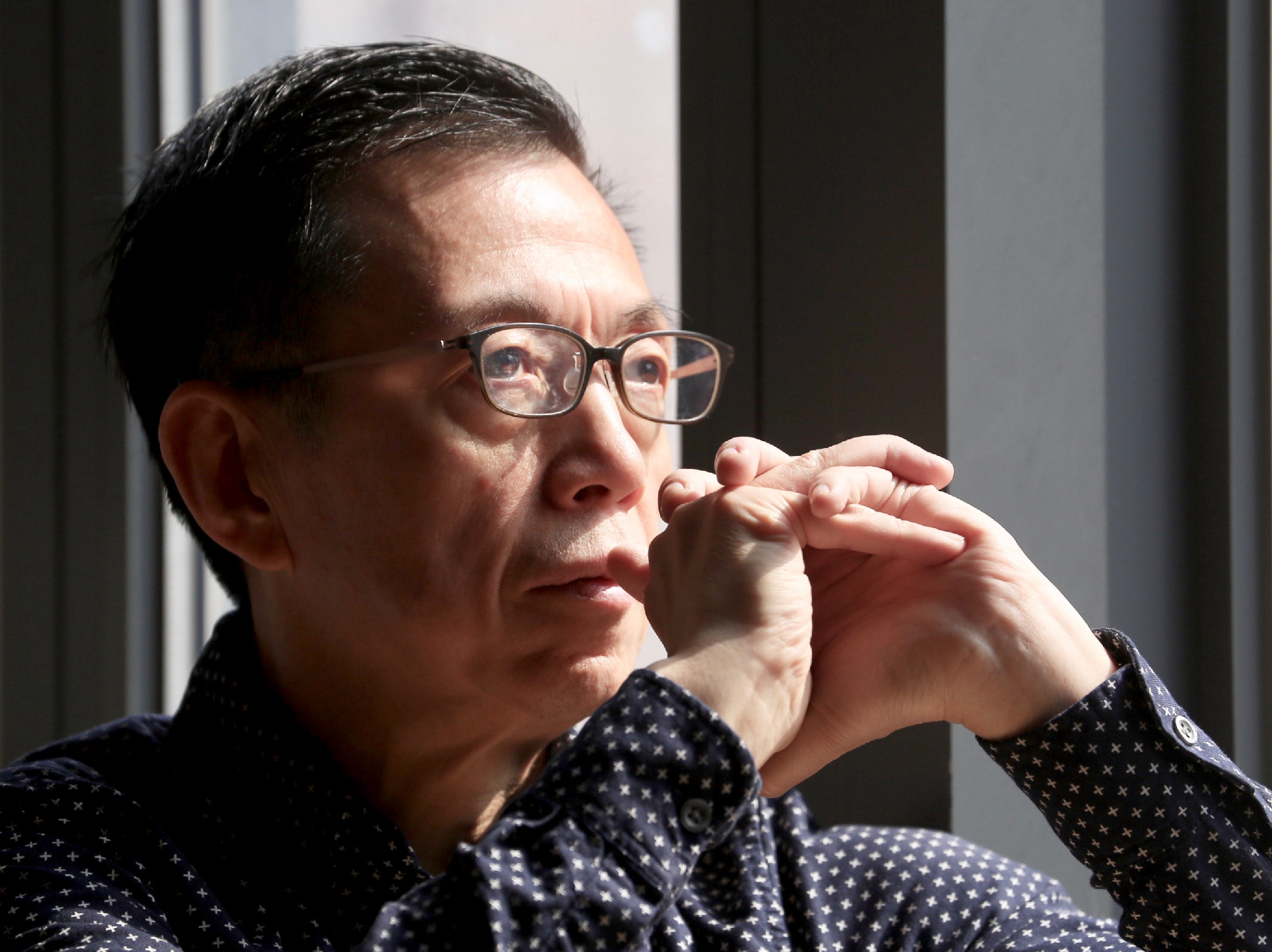 A middle-aged man with glasses and dark hair, wearing a dark shirt with small white dots, sits with his hands clasped near his face in a contemplative pose, near a window with sunlight illuminating part of his face.