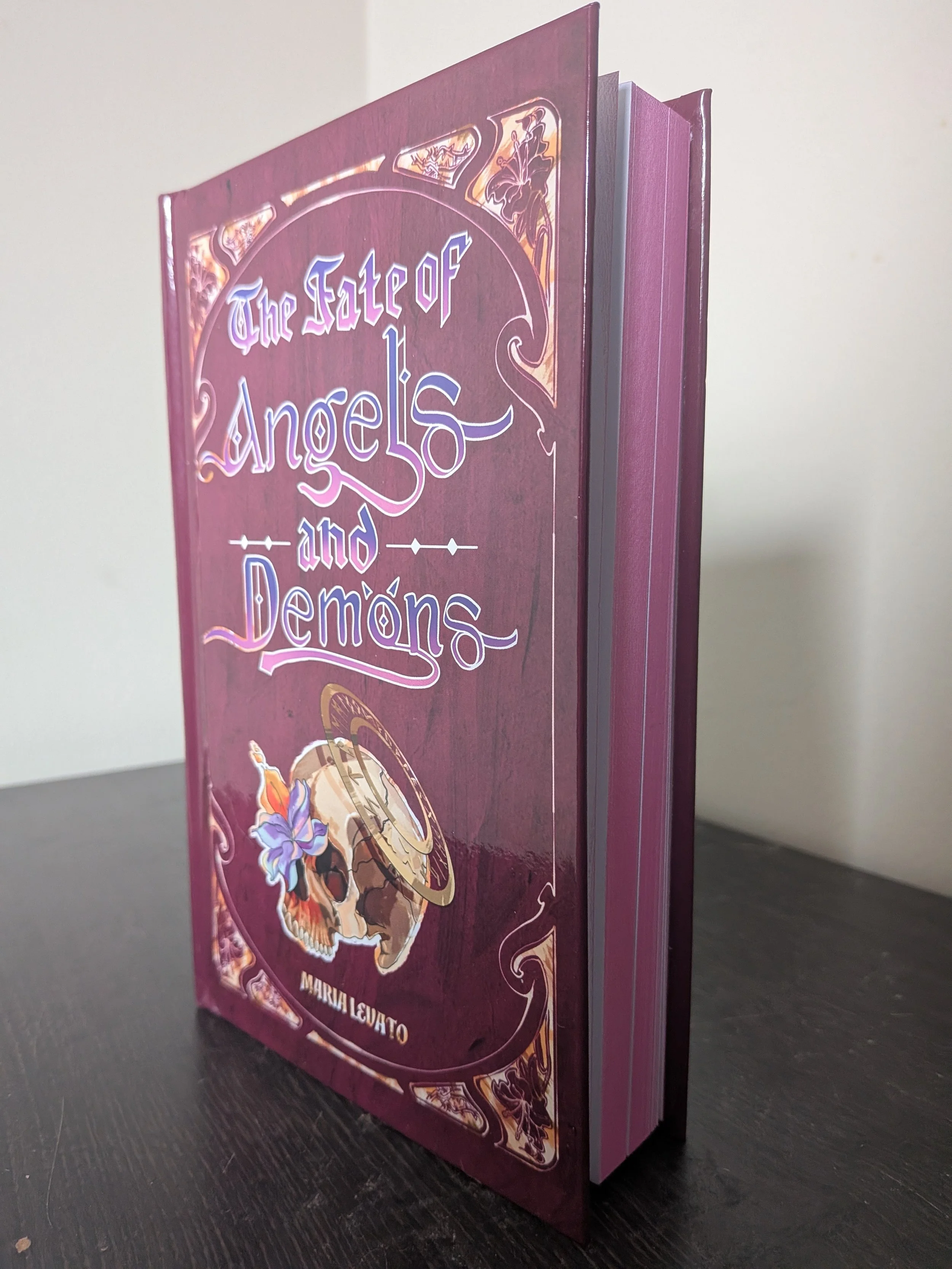 The Fate of Angels and Demons by Maria Levato (special edition hardcover). The angle shows the skull and title on the front cover while also catching the pink sprayed edges.