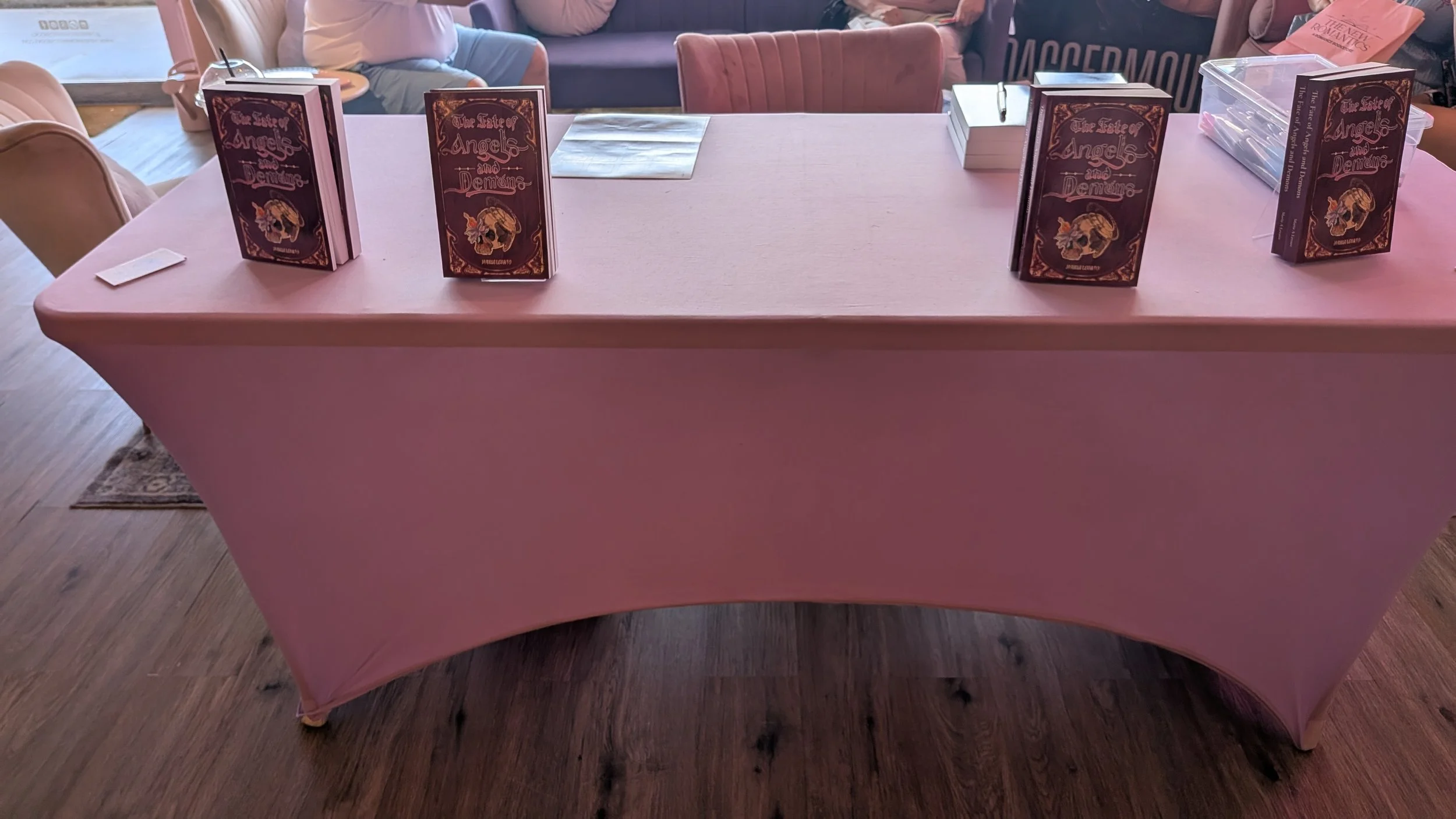 A pink table with books titled 'The Tale of Angels and Demons' arranged upright on it, in a room with people sitting on sofas in the background.